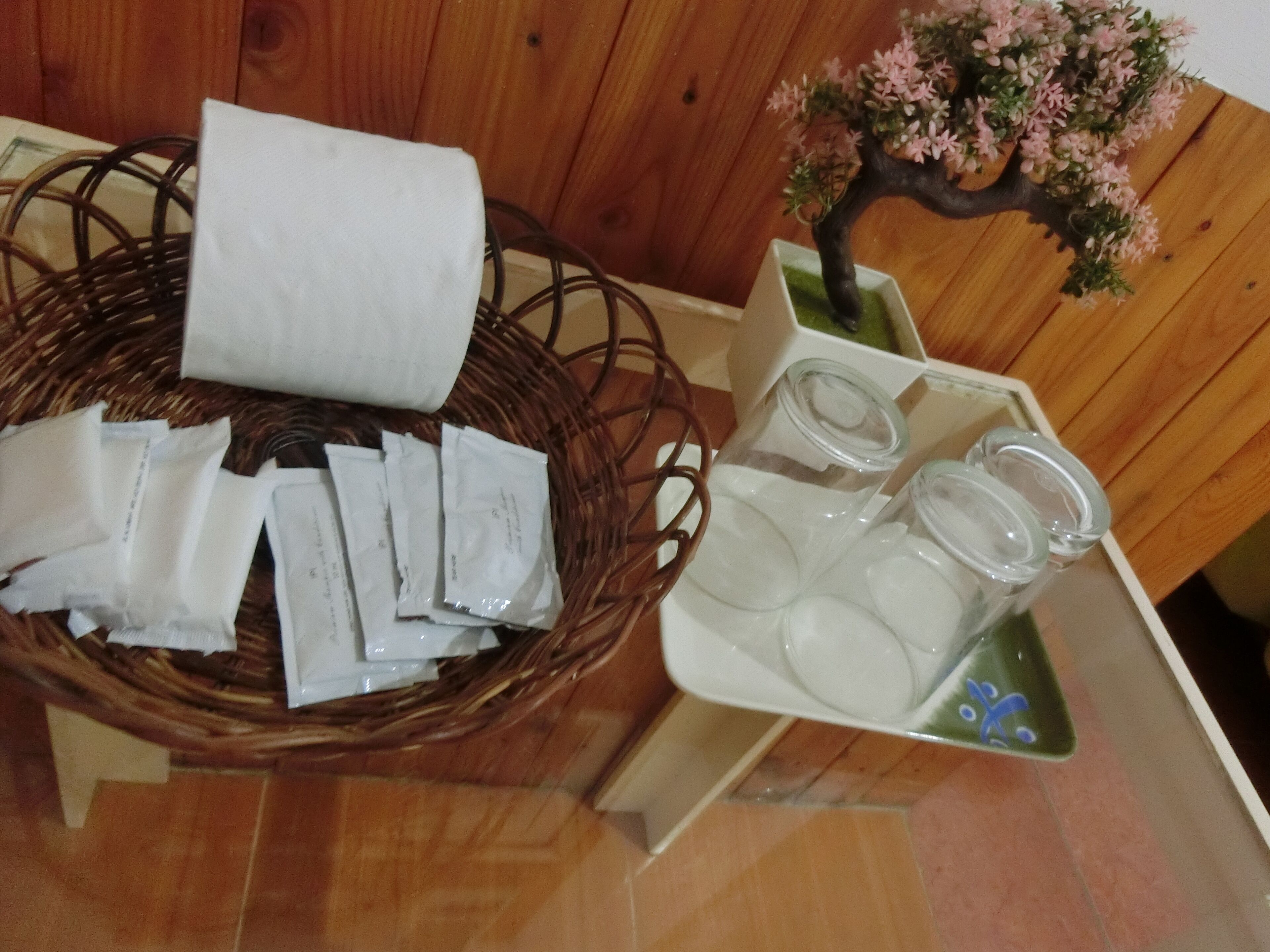 Una cesta de mimbre con papel de seda y pequeños paquetes, un tarro de cristal con tapas, y una pequeña planta en maceta con flores rosas.