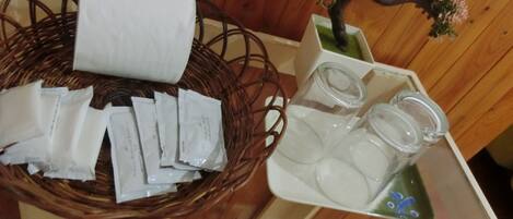 Una cesta de mimbre con papel de seda y pequeños paquetes, un tarro de cristal con tapas, y una pequeña planta en maceta con flores rosas.