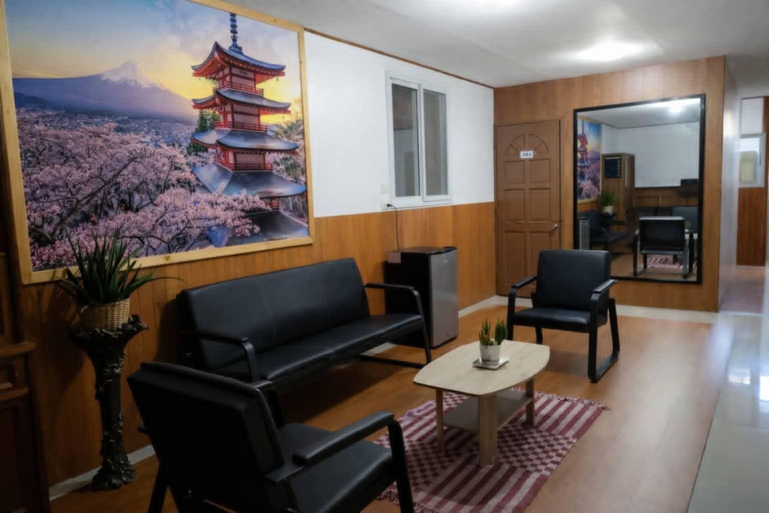 A room with a sofa, chairs, a small table, a potted plant, a painting of a pagoda, and a door with a sign.