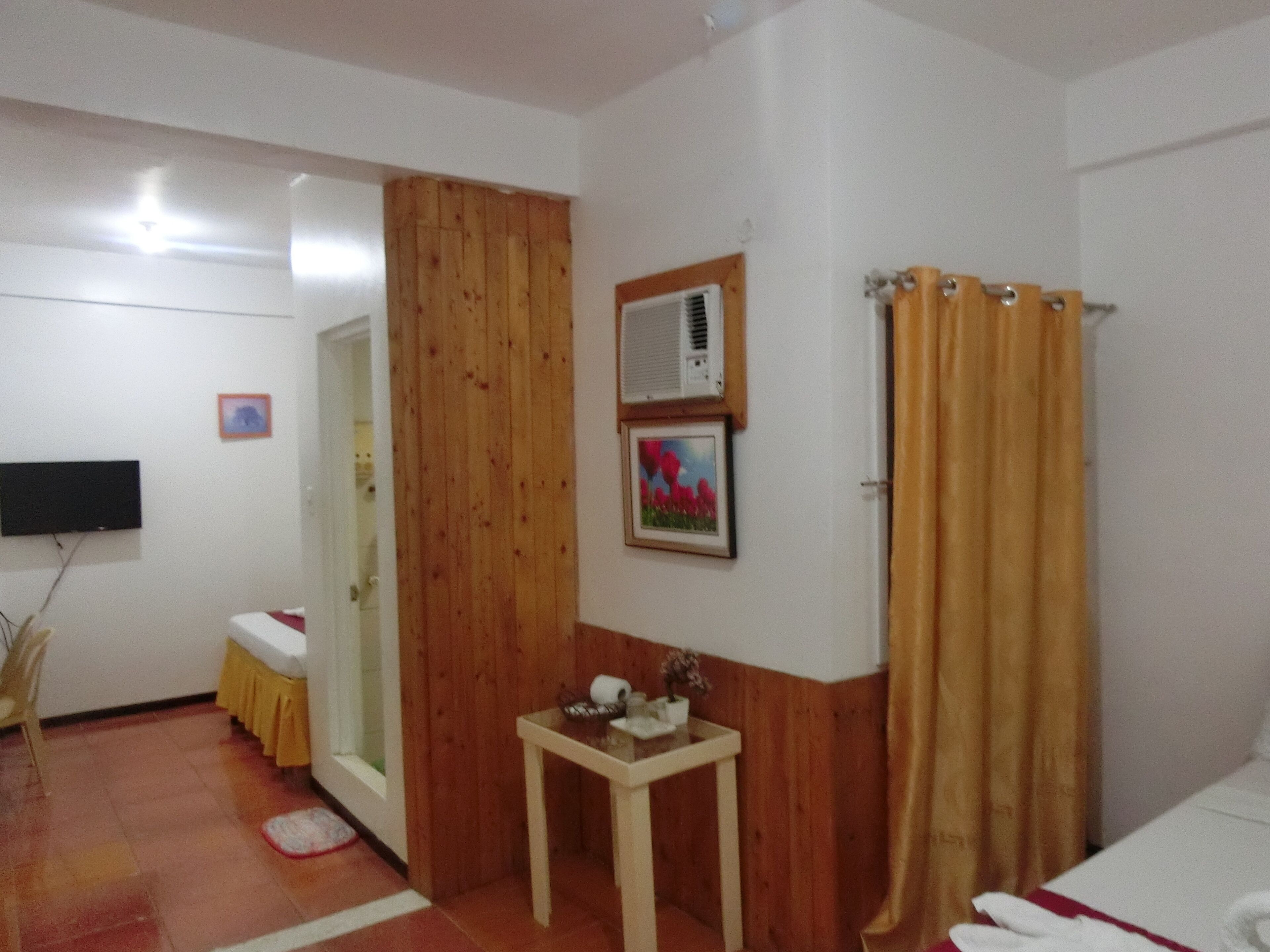 Habitación de hotel con puerta de madera, una mesita con un florero, un televisor de pantalla plana en la pared y una cortina de ducha.