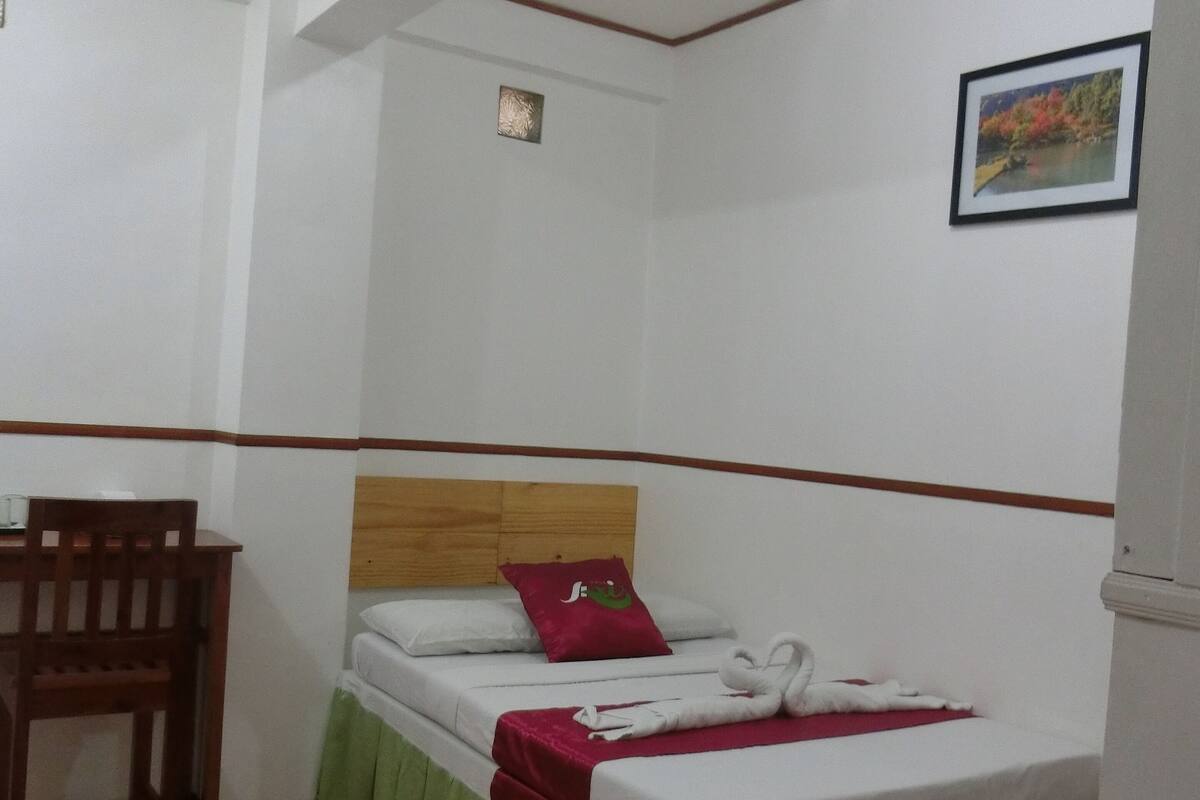 A small, clean room with a single bed, a wooden chair, and a desk.