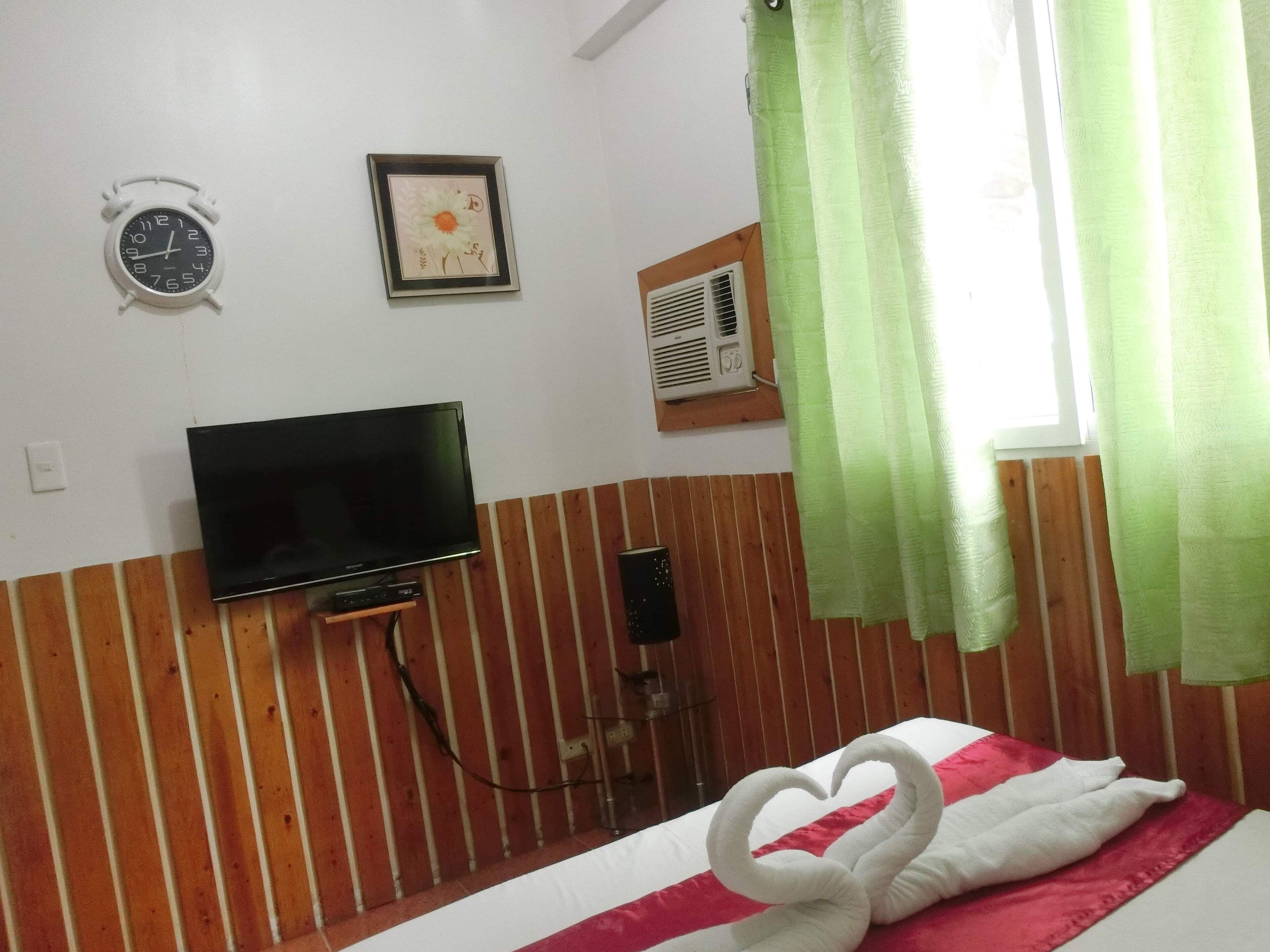 Une chambre d’hôtel avec un lit, un téléviseur fixé au mur, une horloge et une fenêtre avec des rideaux verts.