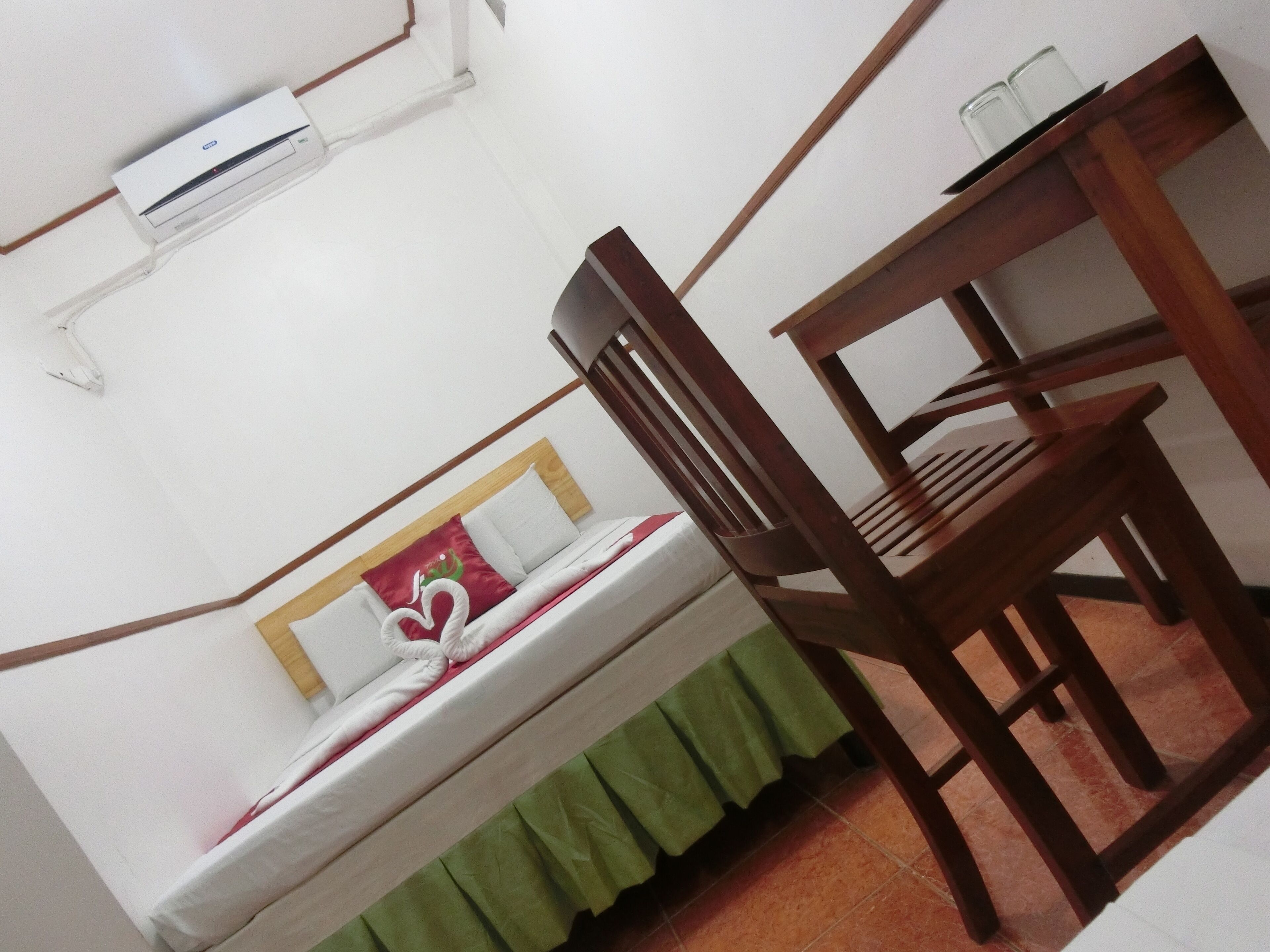 A room with a bed, a wooden chair, a table with two glasses, and an air conditioning unit on the wall.