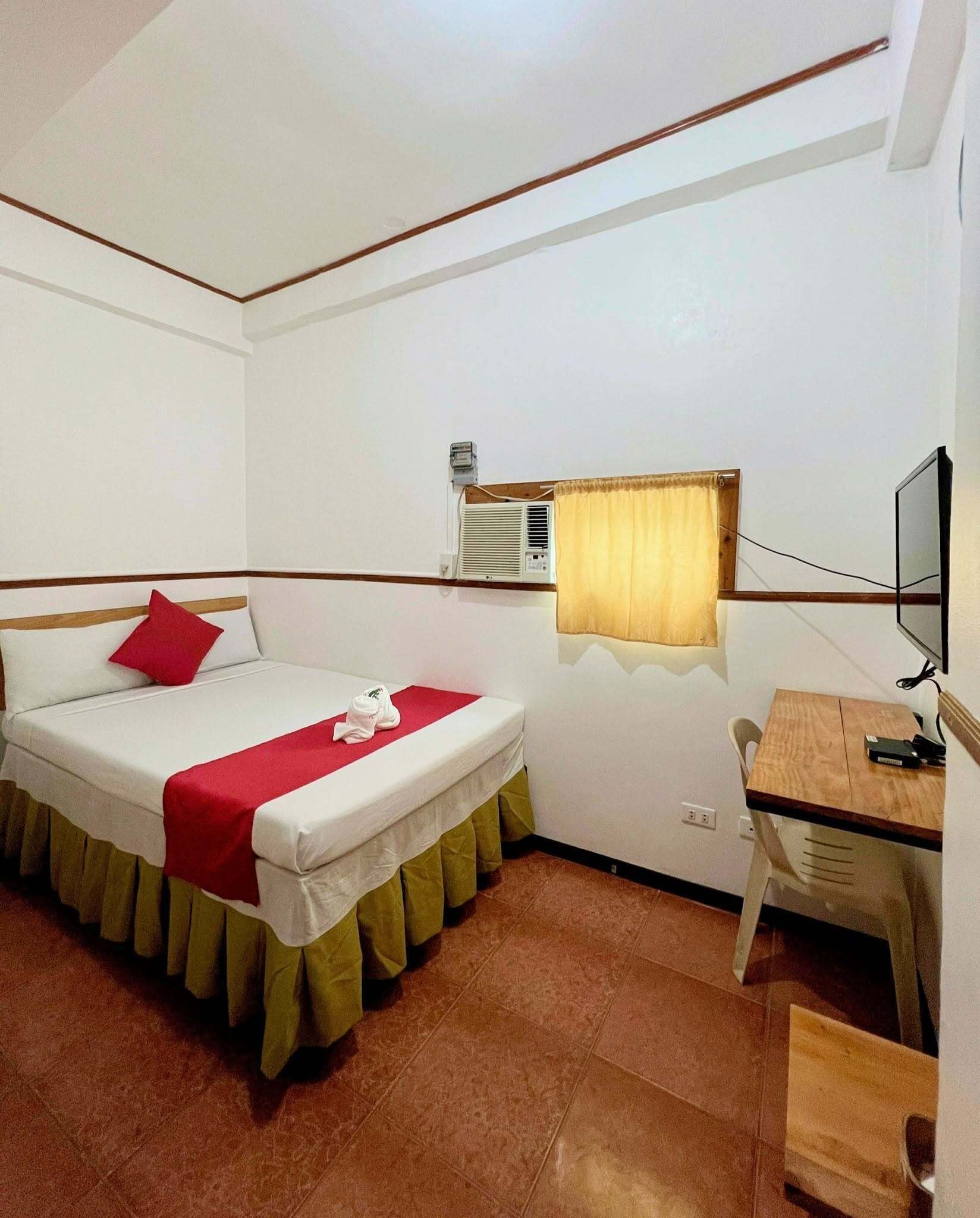 A single bed with a red pillow, a wooden desk with a chair, a television, and a window with a curtain.