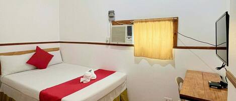 A single bed with a red pillow, a wooden desk with a chair, a television, and a window with a curtain.