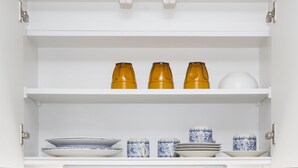 A white cabinet with three shelves. The top shelf has three brown glasses and a white bowl. The middle shelf has three blue and white cups and a white bowl. The bottom shelf has a white plate, a blue and white cup, and a white bowl. - JESUS SQUARE Nippombashi (Osaka)