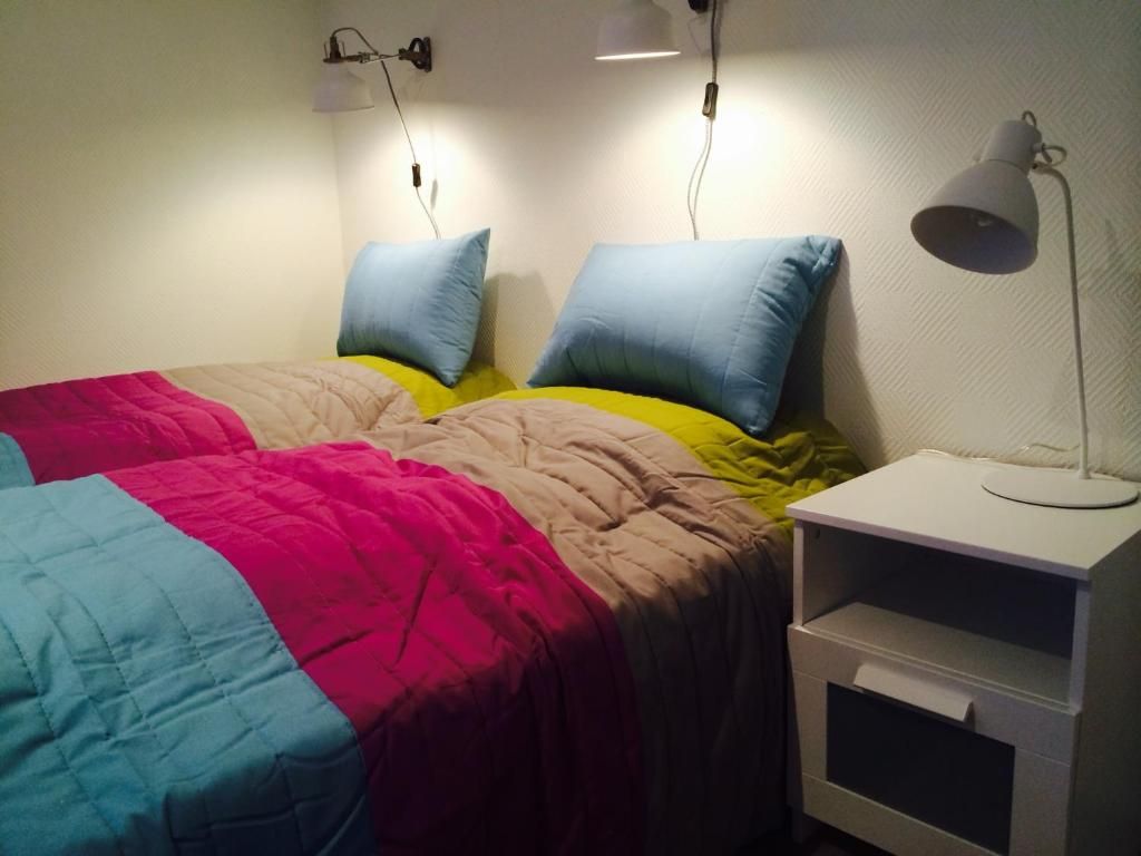 A bedroom with two beds, each with a different colored bedspread. There is a bedside table with a lamp and a drawer.