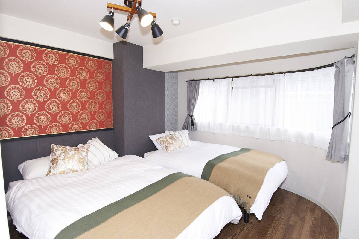 A hotel room with two beds, a red patterned wall, a window with curtains, and a ceiling light fixture.