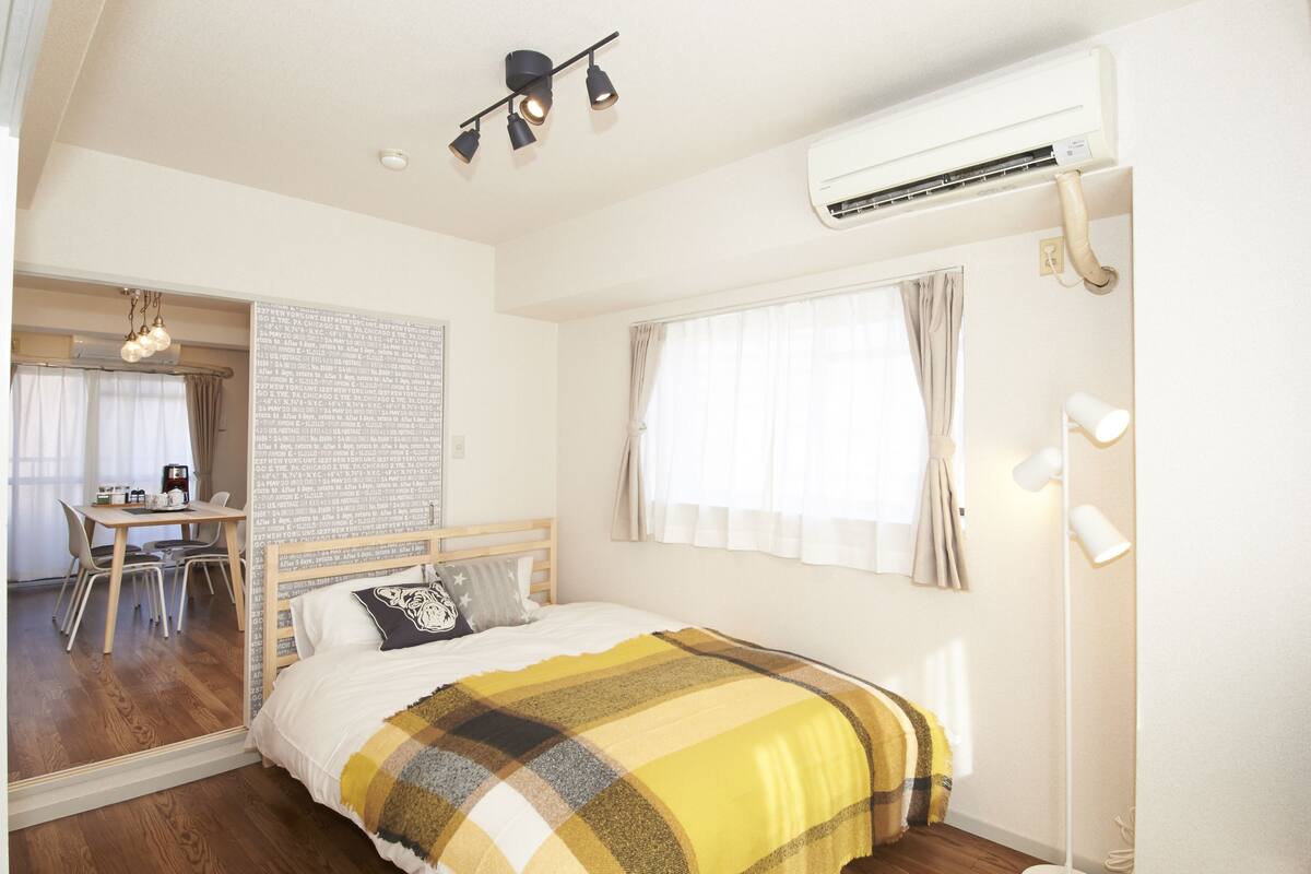 A bedroom with a bed, a desk, and a chair. There is a window with curtains, an air conditioner, and a floor lamp.