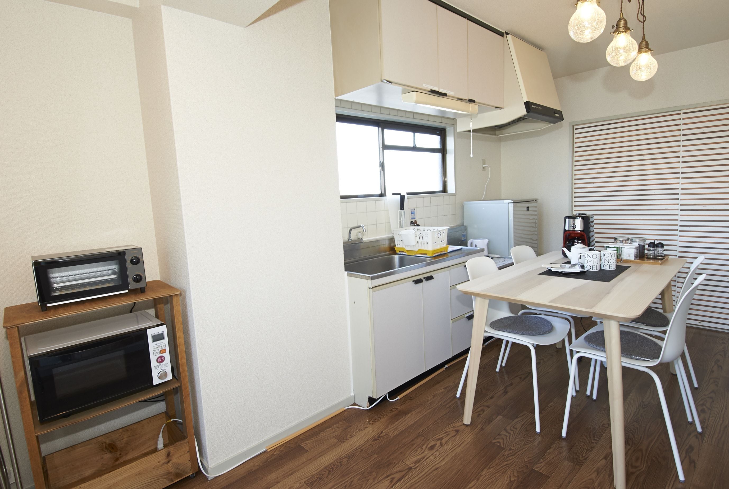 A compact kitchen with a microwave, toaster, and sink. There is a dining table with chairs and a window with blinds.
