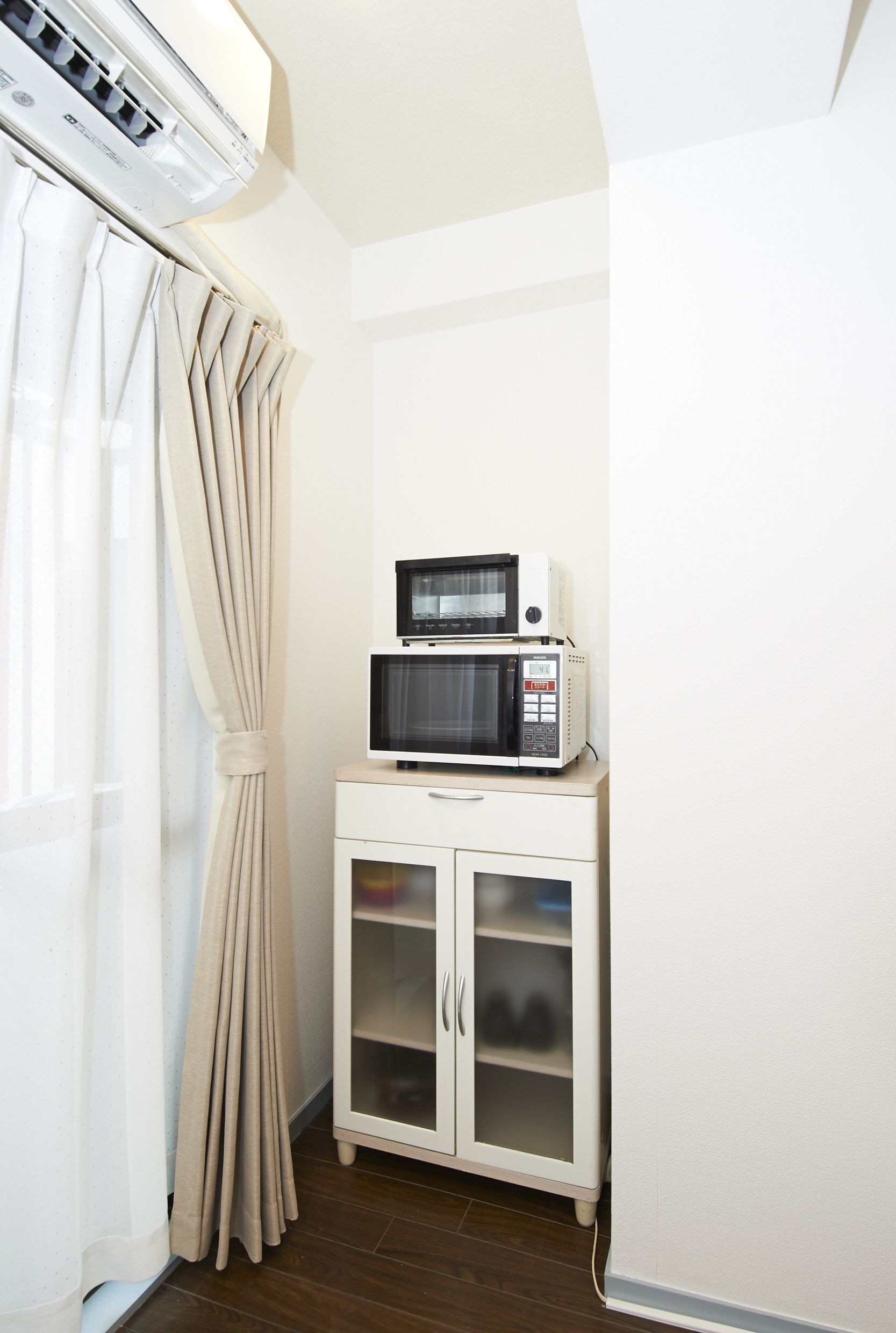 A small kitchenette with a microwave on a cabinet, a white wall, and a curtain.