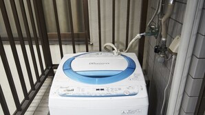 Una lavadora compacta y blanca con tapa azul, ubicada en un espacio reducido con paredes de azulejos y una baranda metálica.