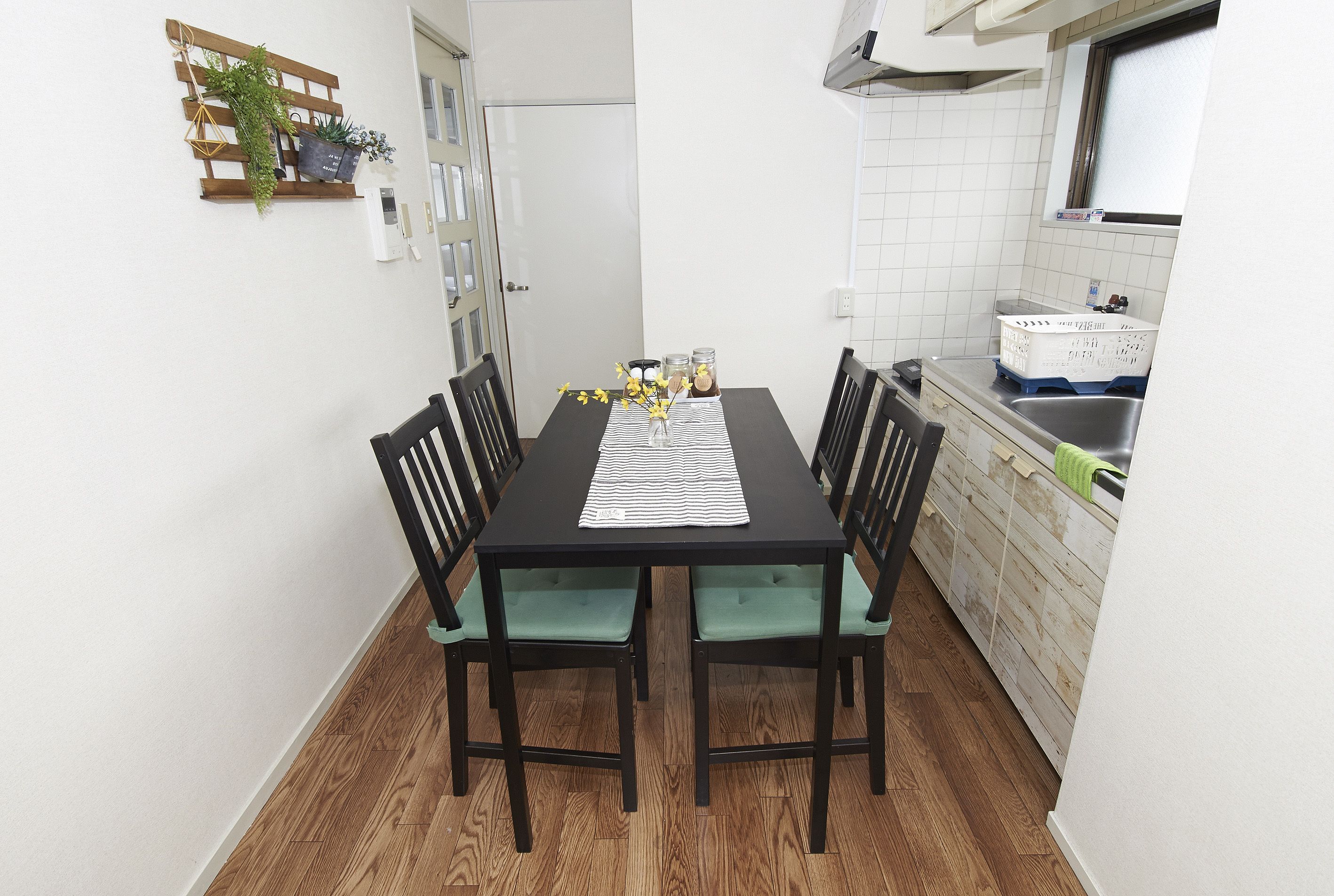 A small dining area with a black table and green cushioned chairs, a wooden floor, and a kitchen with white tiles and a window.