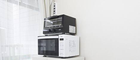 Una cocina con microondas y horno sobre un mueble, un cubo de basura y una ventana con cortina ligera.