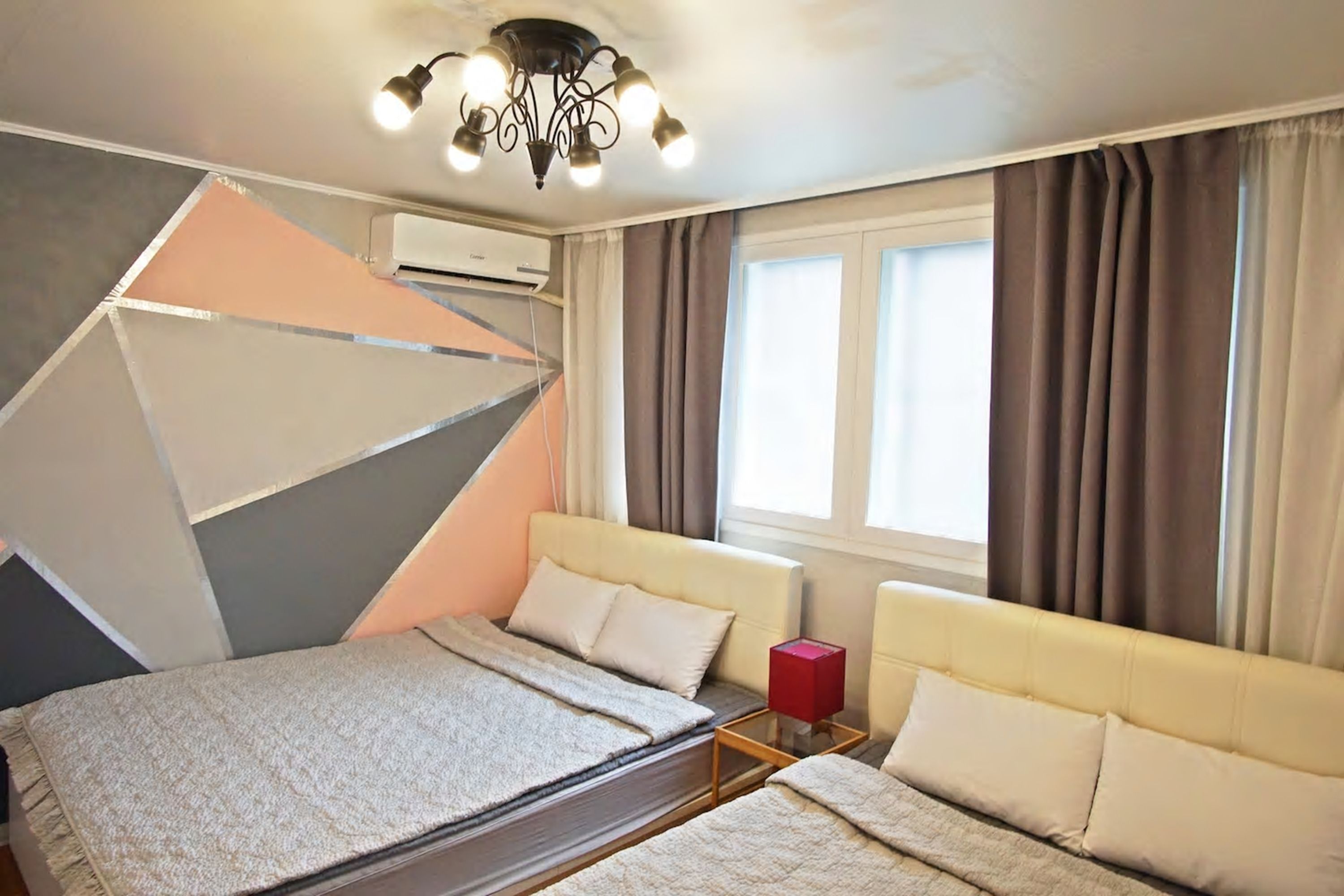 A modern hotel room with a bed, two sofas, a red cube-shaped table, and a window with curtains.