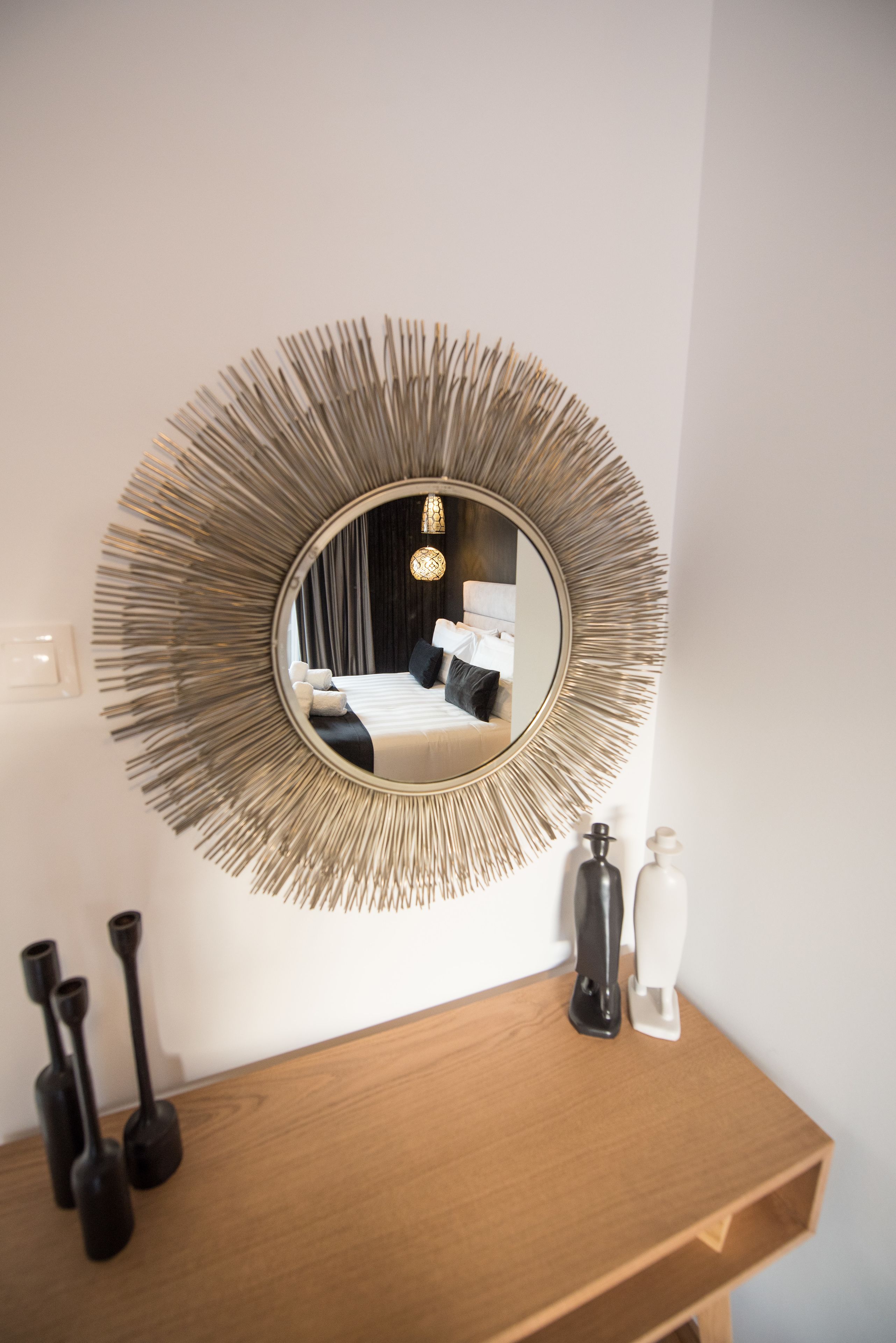 A round mirror with a sunburst frame on a wooden dresser, reflecting a bedroom with a bed and a chandelier.