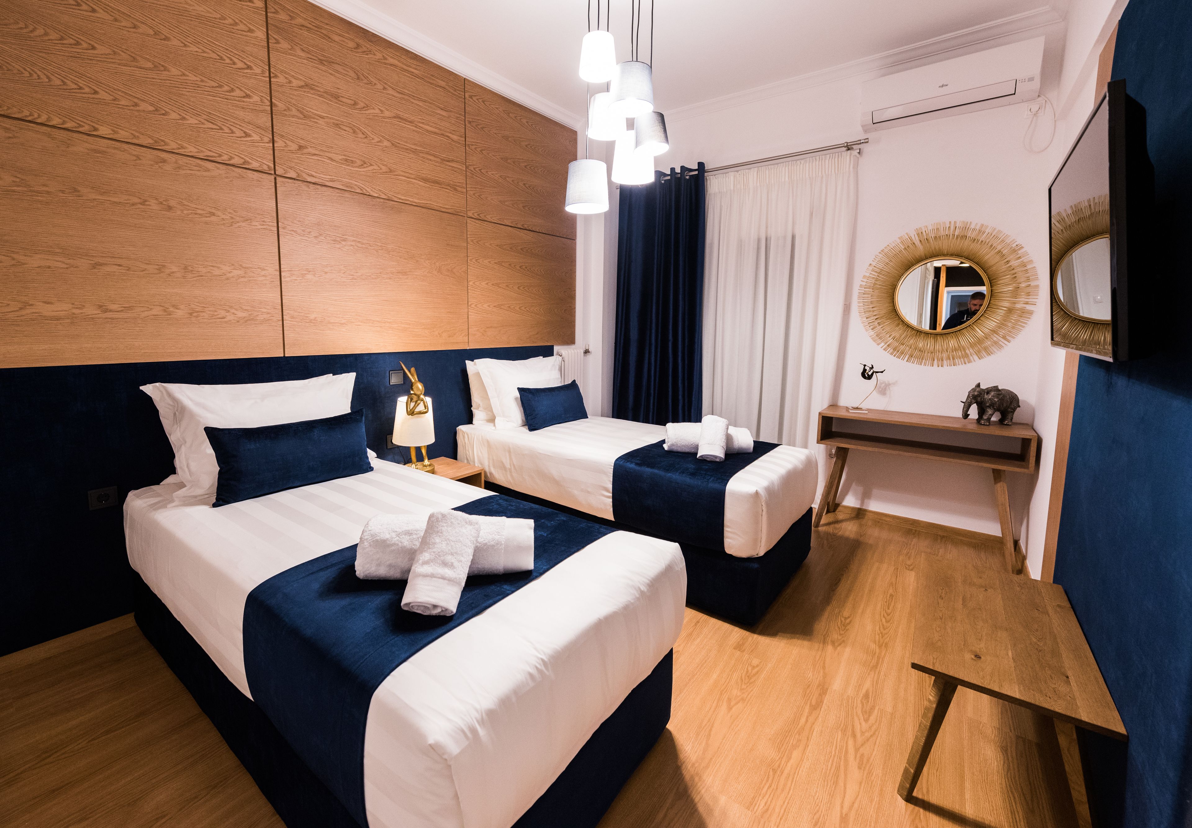 A hotel room with two beds, a wooden headboard, a bedside table with a lamp, a mirror, and a TV.