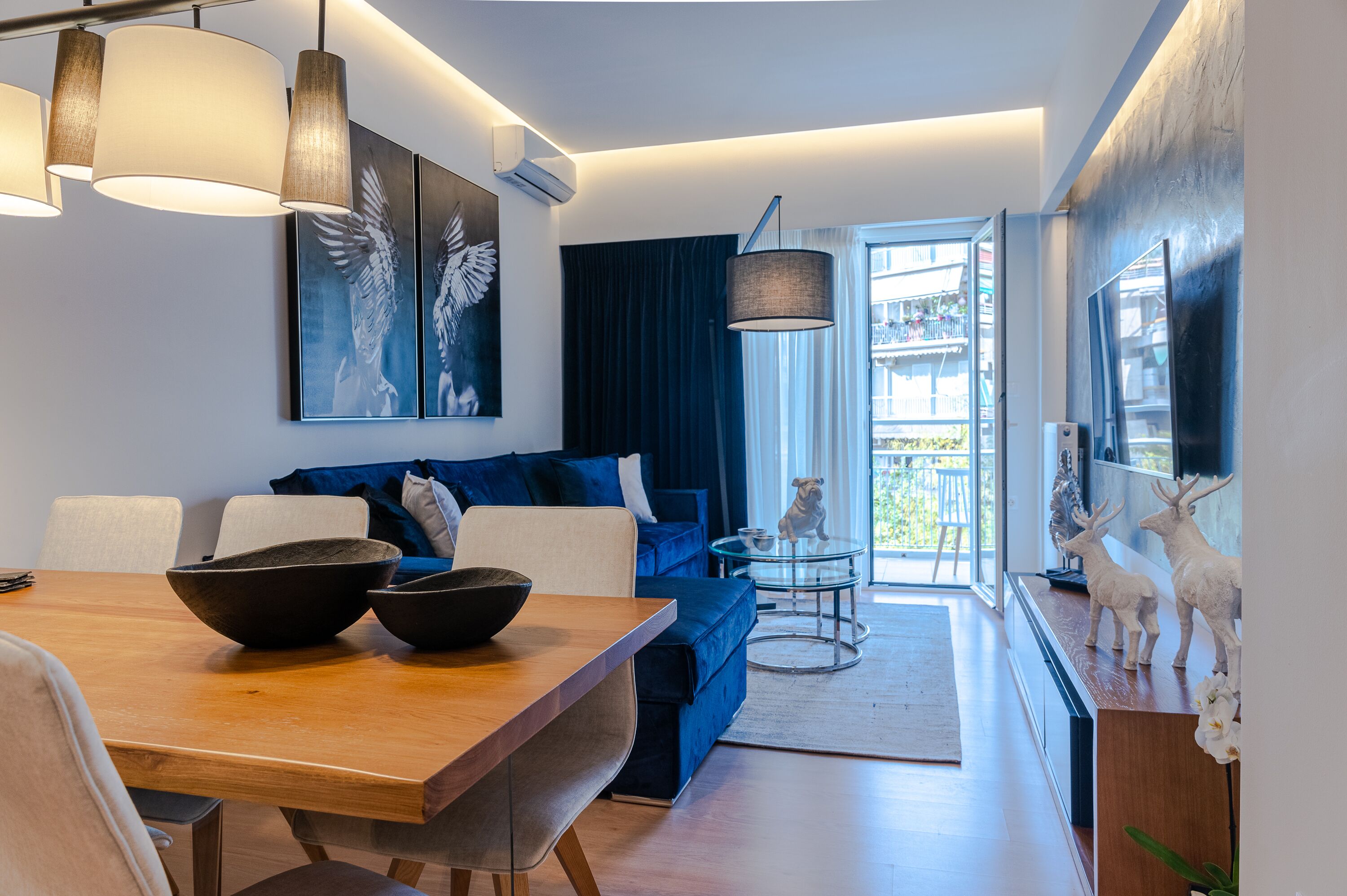 A modern living room with a wooden dining table, blue sofa, and a glass coffee table. There is a balcony with a view of the city.
