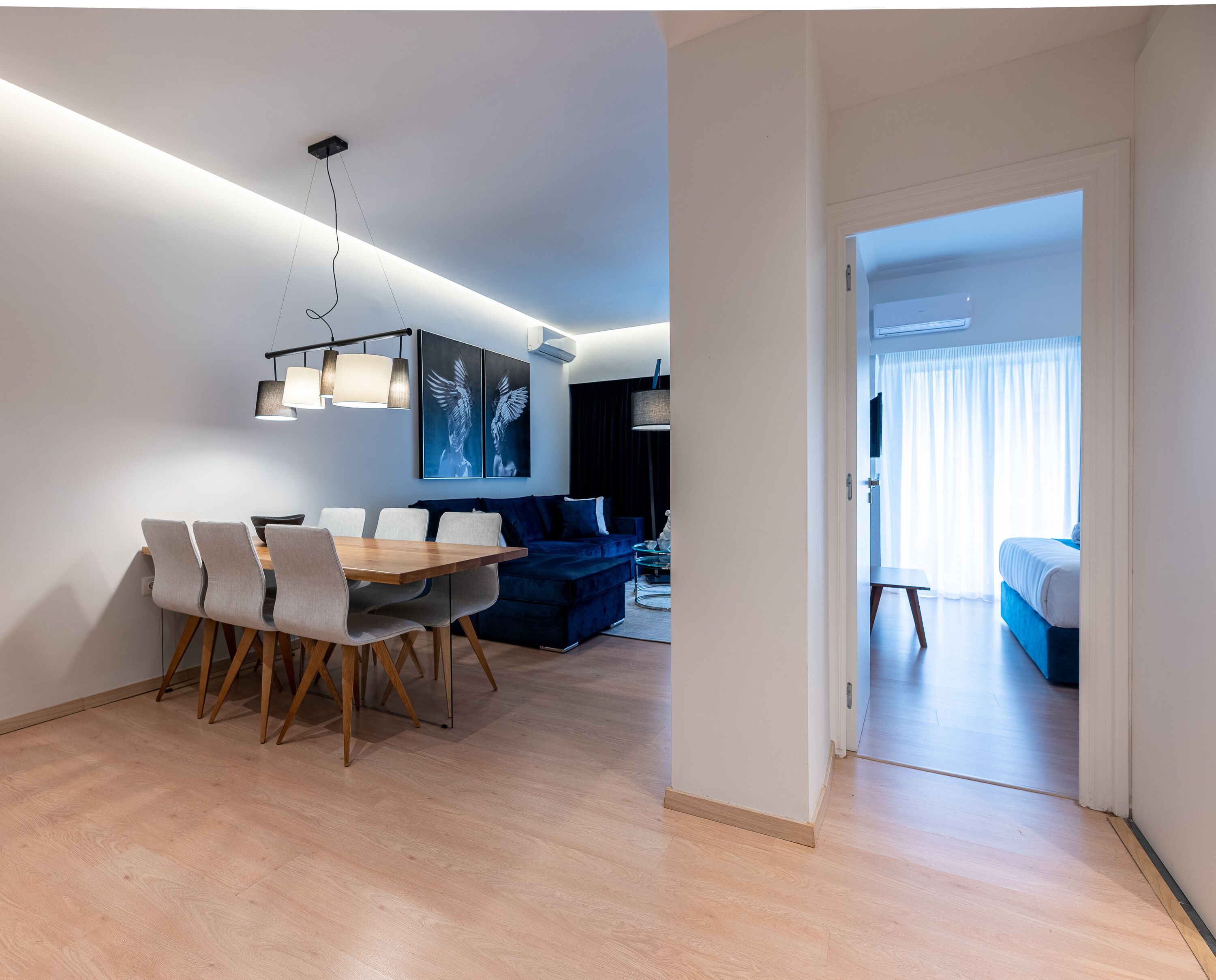 A modern living room with a dining area, featuring a wooden dining table, chairs, a blue sofa, and a large artwork on the wall.