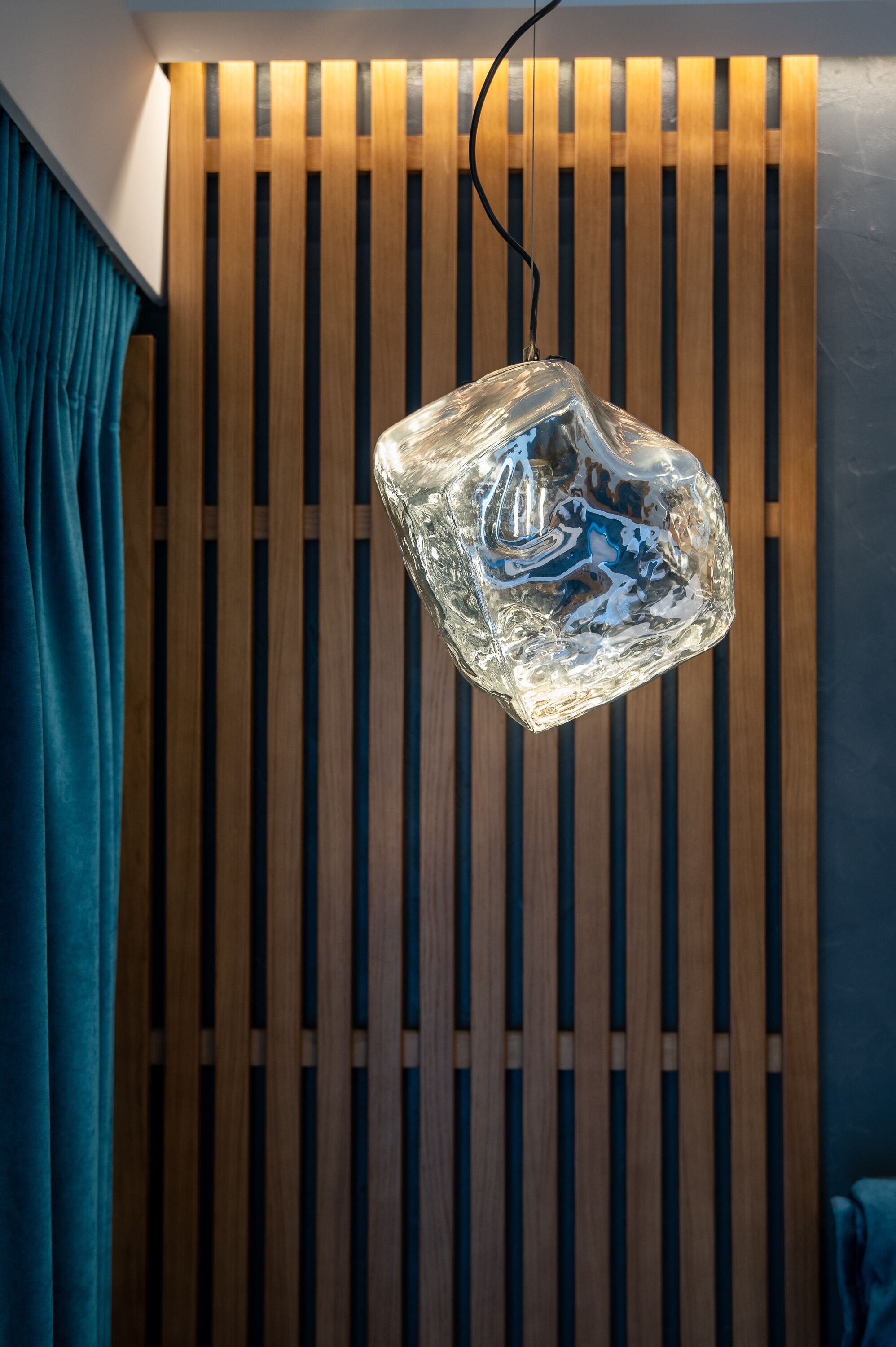 En hængende lampe med geometrisk glasdesign, hængt under en loftbeklædning af trælameller med et blåt gardin i baggrunden.