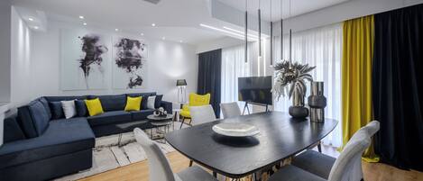 Una sala de estar moderna con una mesa de comedor negra, sillas grises, un sofá negro con cojines amarillos, un televisor y una planta.