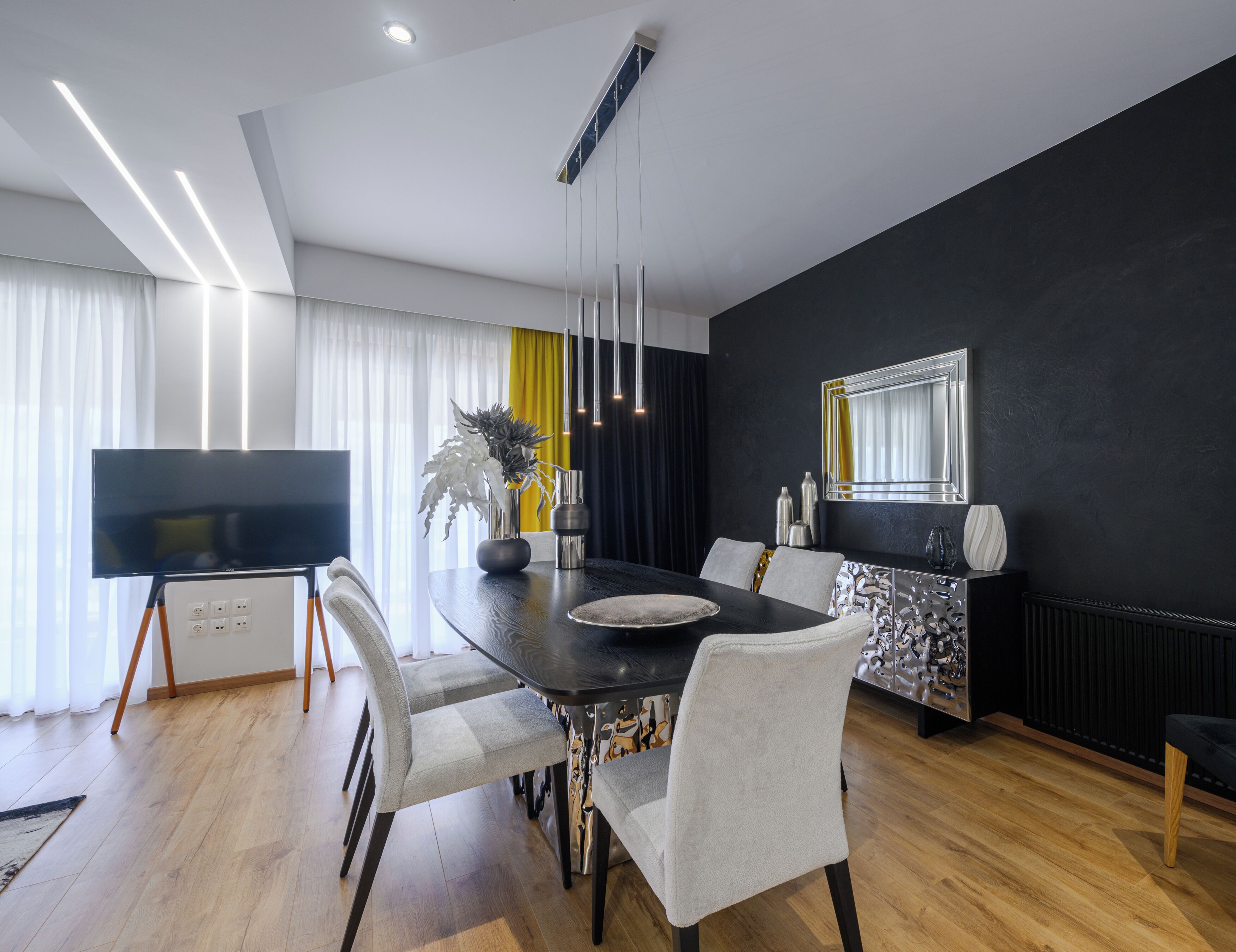 A modern dining area with a dark wooden table, white upholstered chairs, and a large flat-screen TV on a stand.