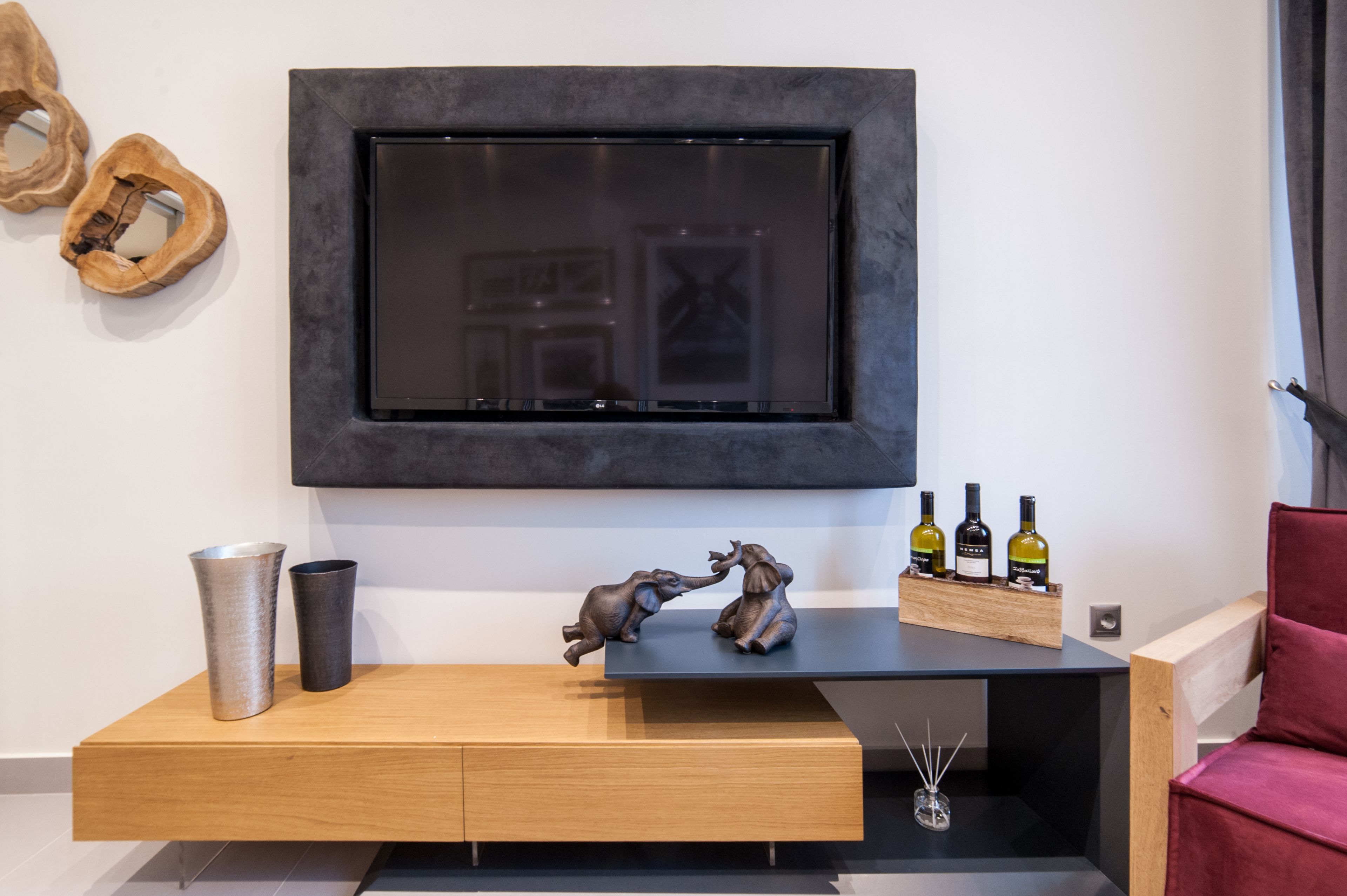Een moderne woonkamer met een wandtelevisie, een houten console, decoratieve objecten en een rode fauteuil.