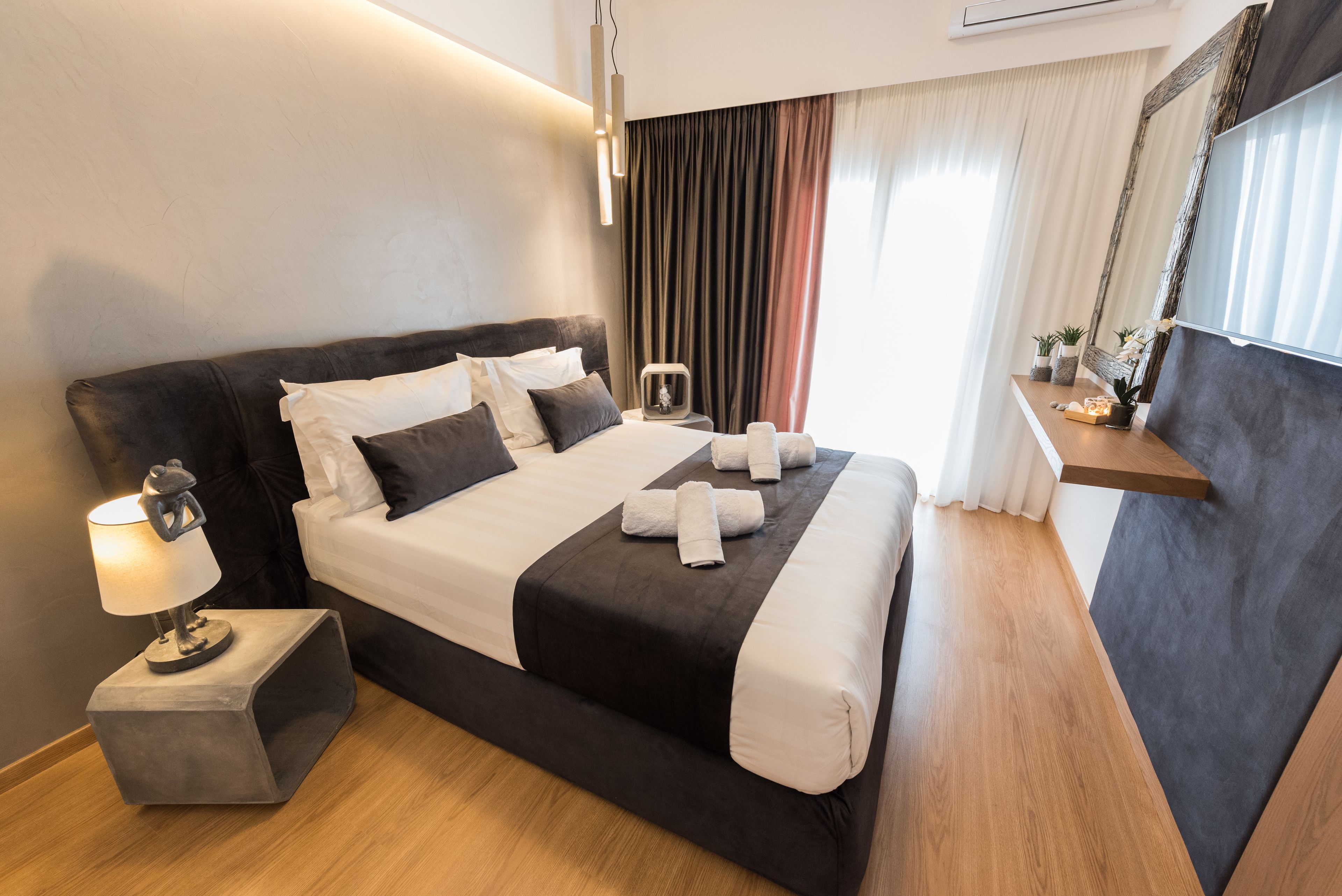 A modern hotel room with a large bed, a bedside table with a lamp, a small side table, and a mirror on the wall.