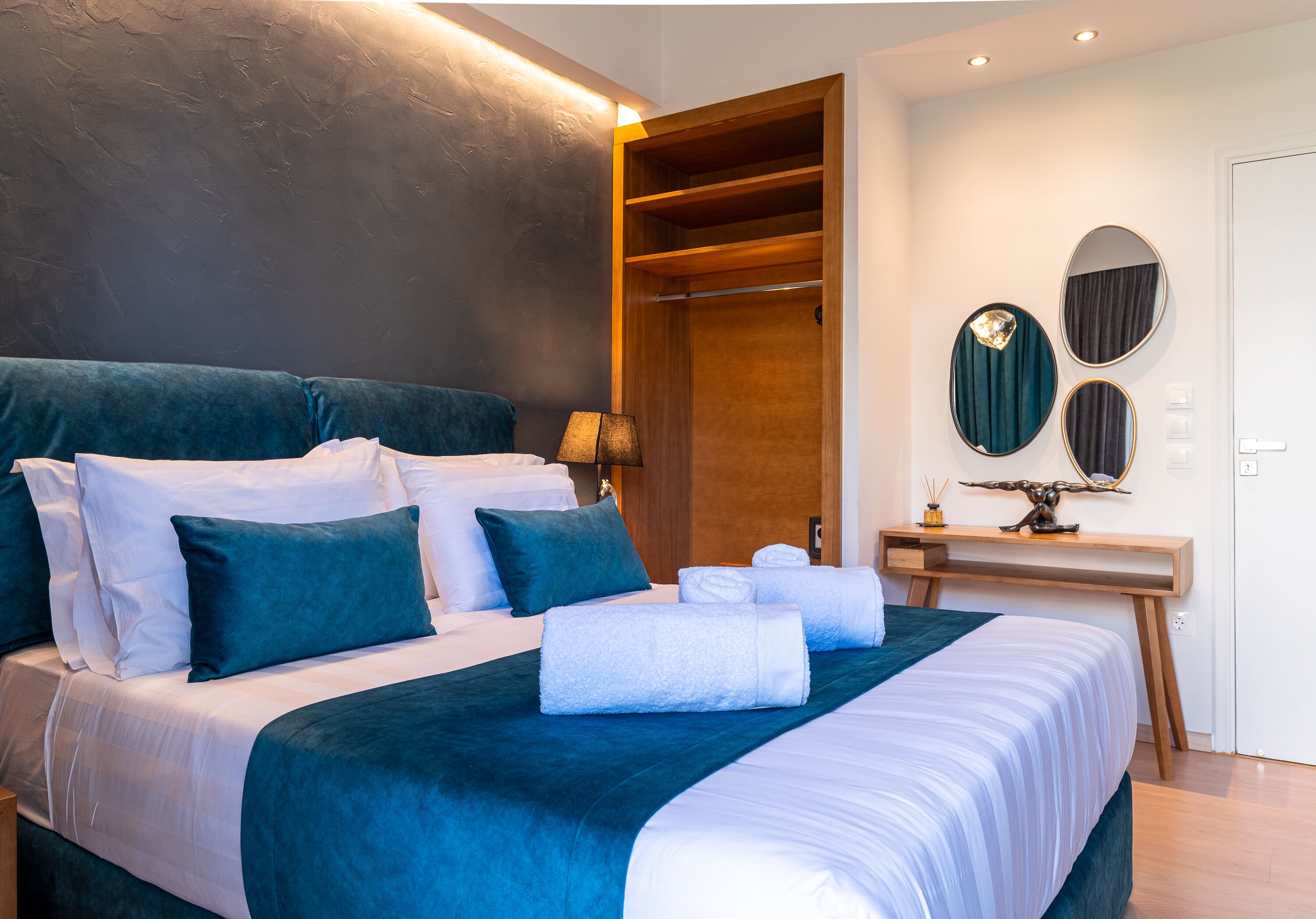 A modern hotel room with a large bed, a wooden wardrobe, a desk with a mirror, and a wall-mounted light fixture.