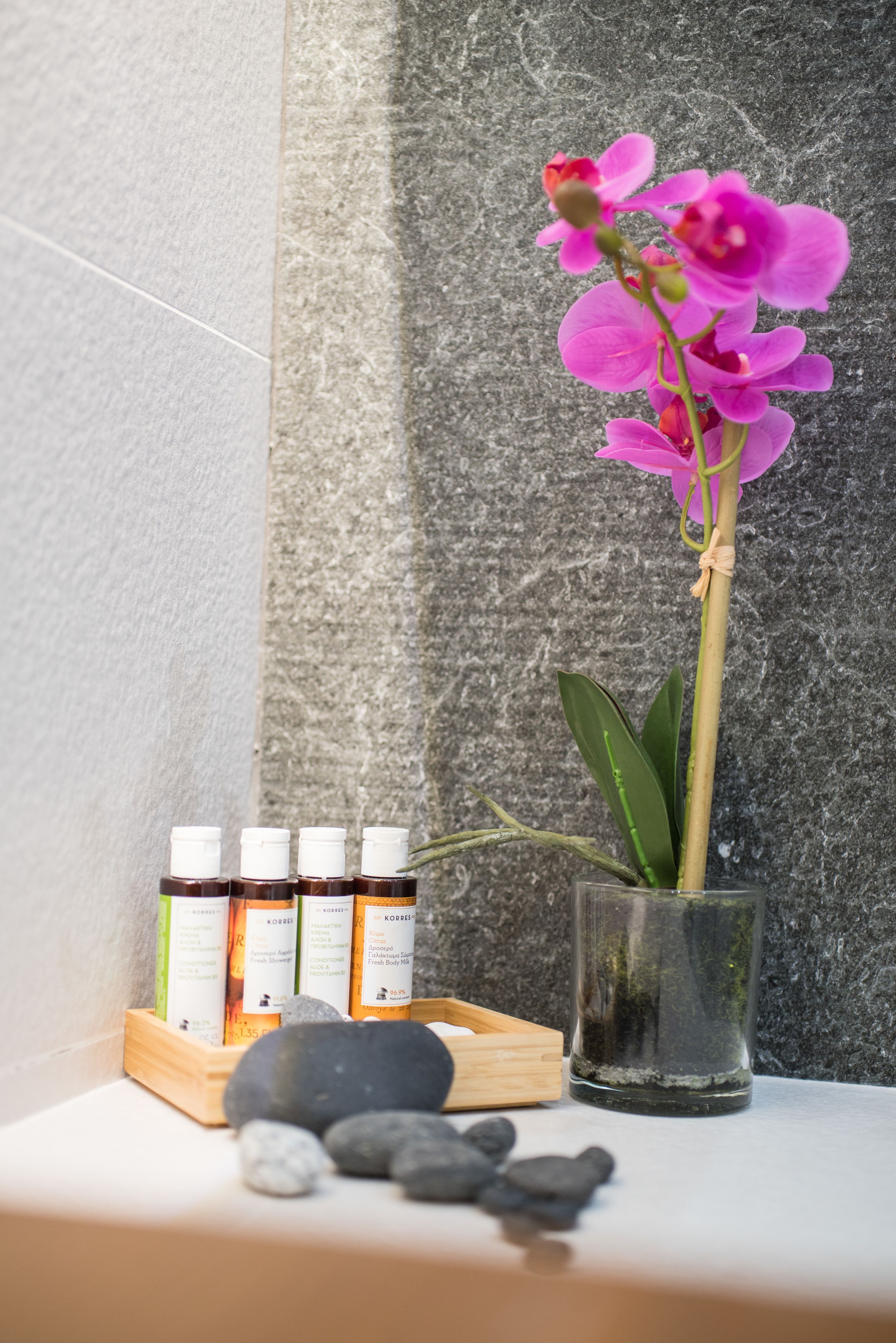 A glass vase with a pink orchid, a wooden tray with bottles, and stones on a surface.