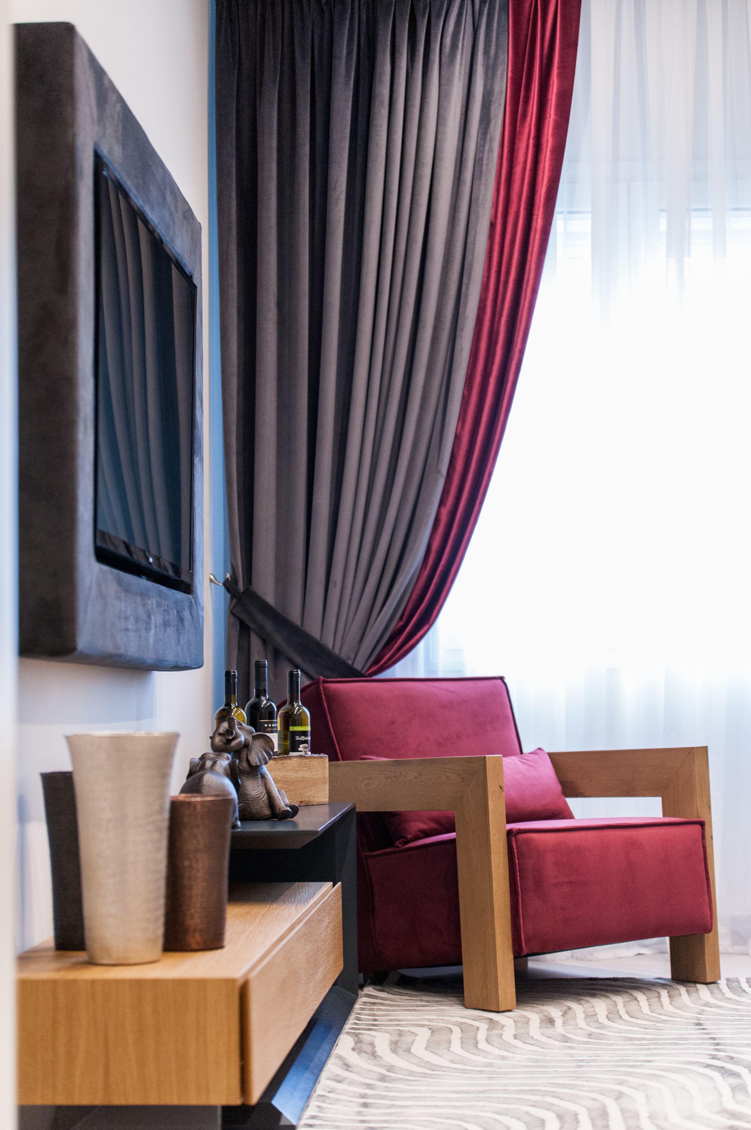 A modern living room with a wooden sofa, a side table with decorative items, and a large window with curtains.