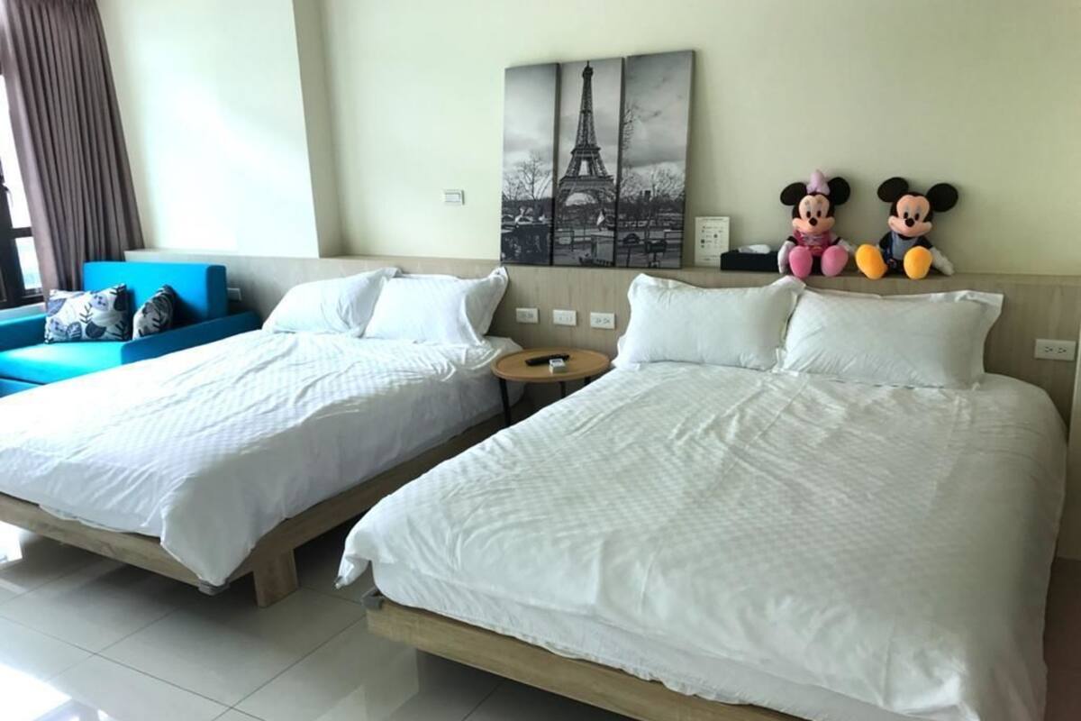 Two single beds with white linens, a blue sofa, a Mickey Mouse and Minnie Mouse plush toy, and a painting of the Eiffel Tower.