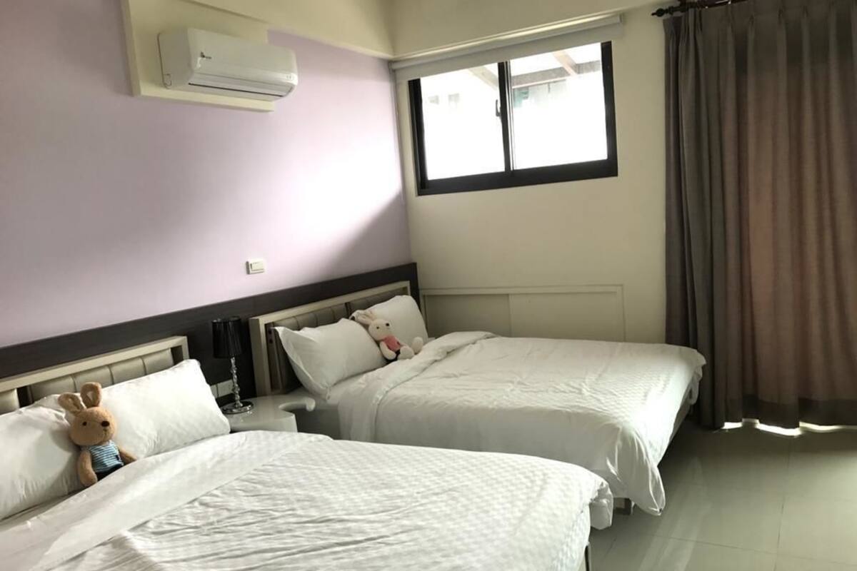A hotel room with two beds, a window with curtains, an air conditioning unit, and a teddy bear on one bed.