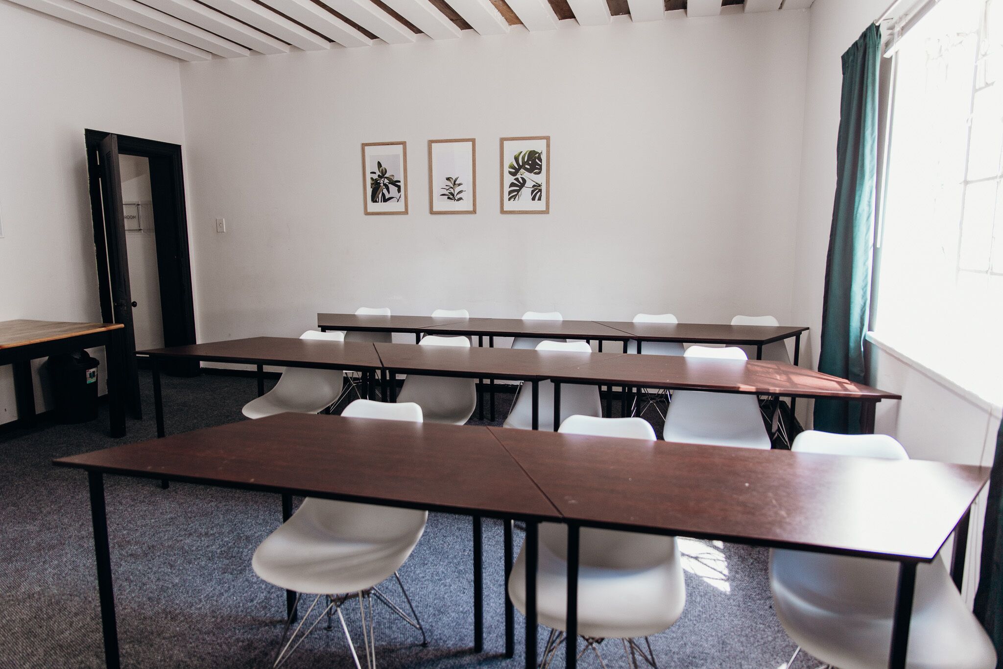 A conference room with multiple tables and chairs, framed artwork on the wall, and a window with curtains.
