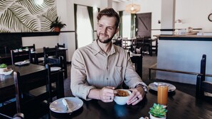 A man dining in a modern restaurant with wooden tables and chairs, a decorative wall, and a bar area in the background. - George Lodge International (George)