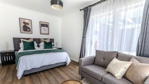 A bedroom with a bed, a grey sofa, wooden side tables, and a large window with sheer curtains. - George Lodge International (George)