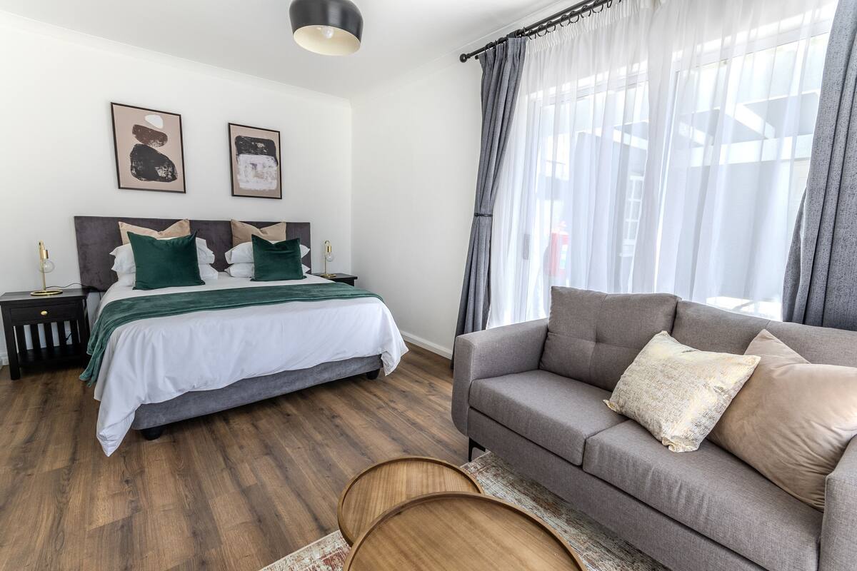 A bedroom with a bed, a grey sofa, wooden side tables, and a large window with sheer curtains.