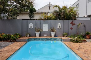 A swimming pool with a tiled edge, surrounded by potted plants and a grey wall with decorative elements. - George Lodge International (George)