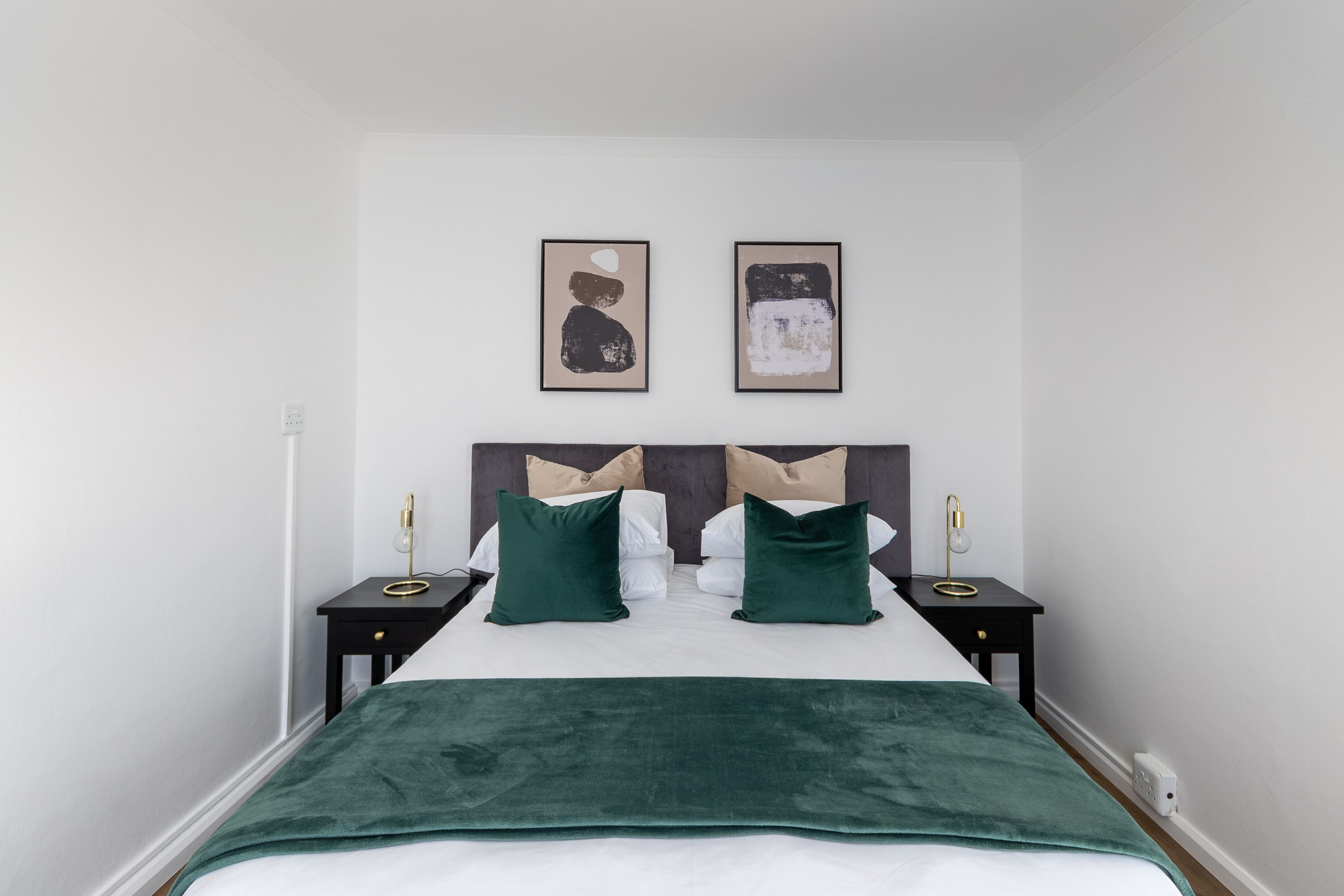 Uma cama com um cobertor verde, duas almofadas e uma cabeceira. Há duas mesas de cabeceira com abajures.