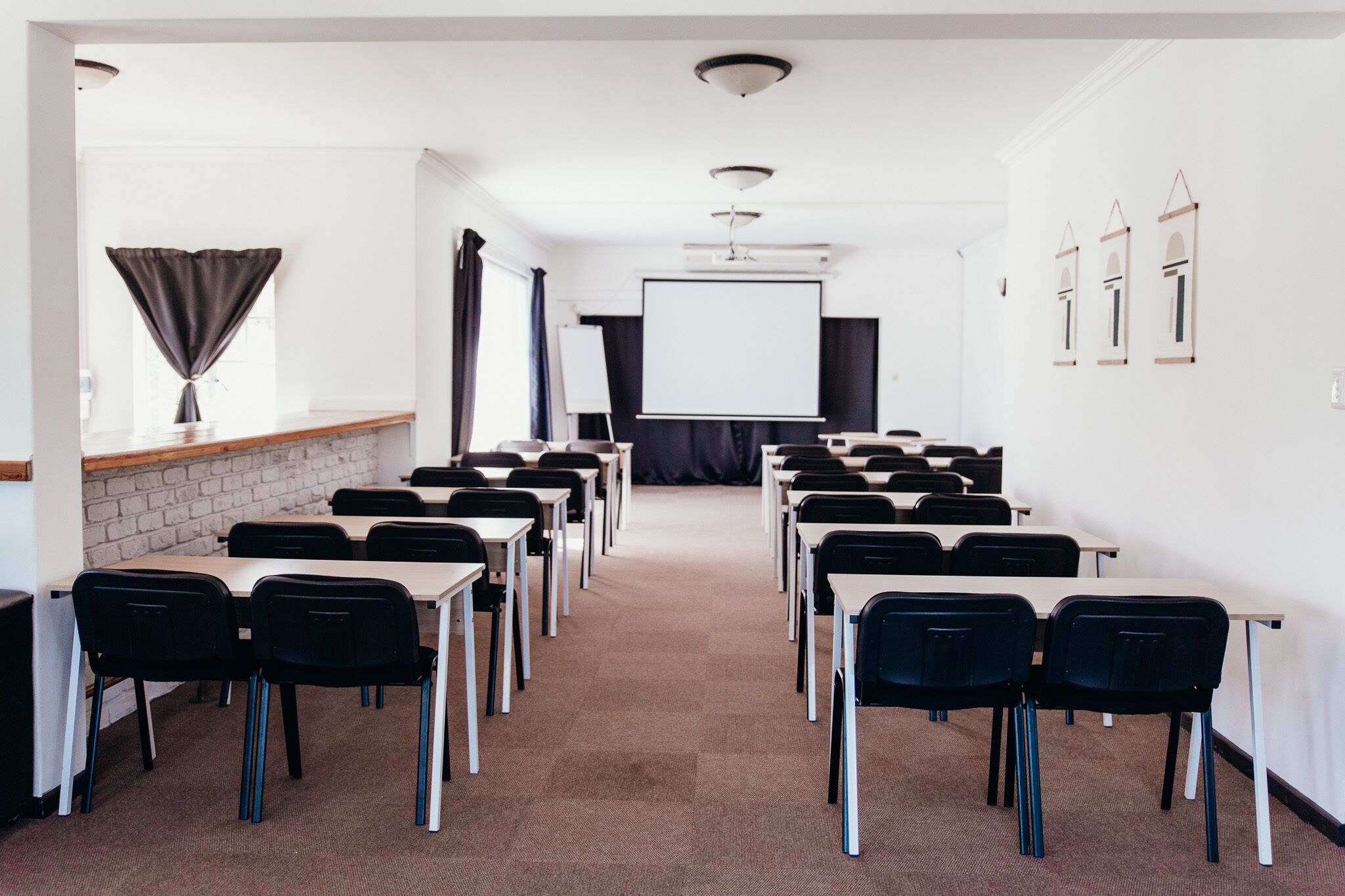 Uma sala de conferências com fileiras de mesas e cadeiras, uma tela de projetor e um quadro branco.