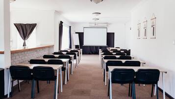 Uma sala de conferências com fileiras de mesas e cadeiras, uma tela de projetor e um quadro branco.