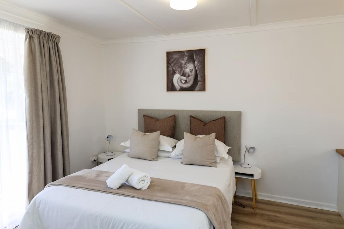 A neatly made bed with a beige blanket, two pillows, and a rolled towel. There is a nightstand with a lamp and a small table beside the bed. A framed picture hangs on the wall above the bed.