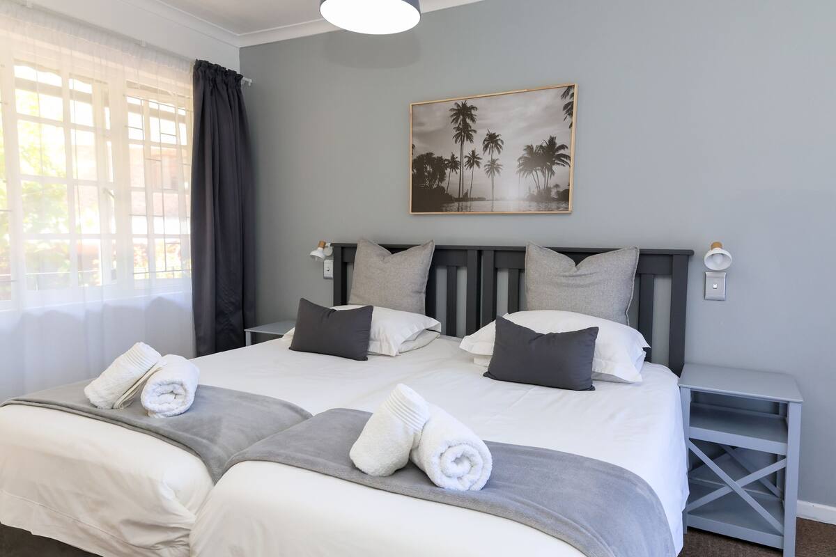 A double bed with two pillows, a bedside table, a nightstand, and a painting of palm trees.