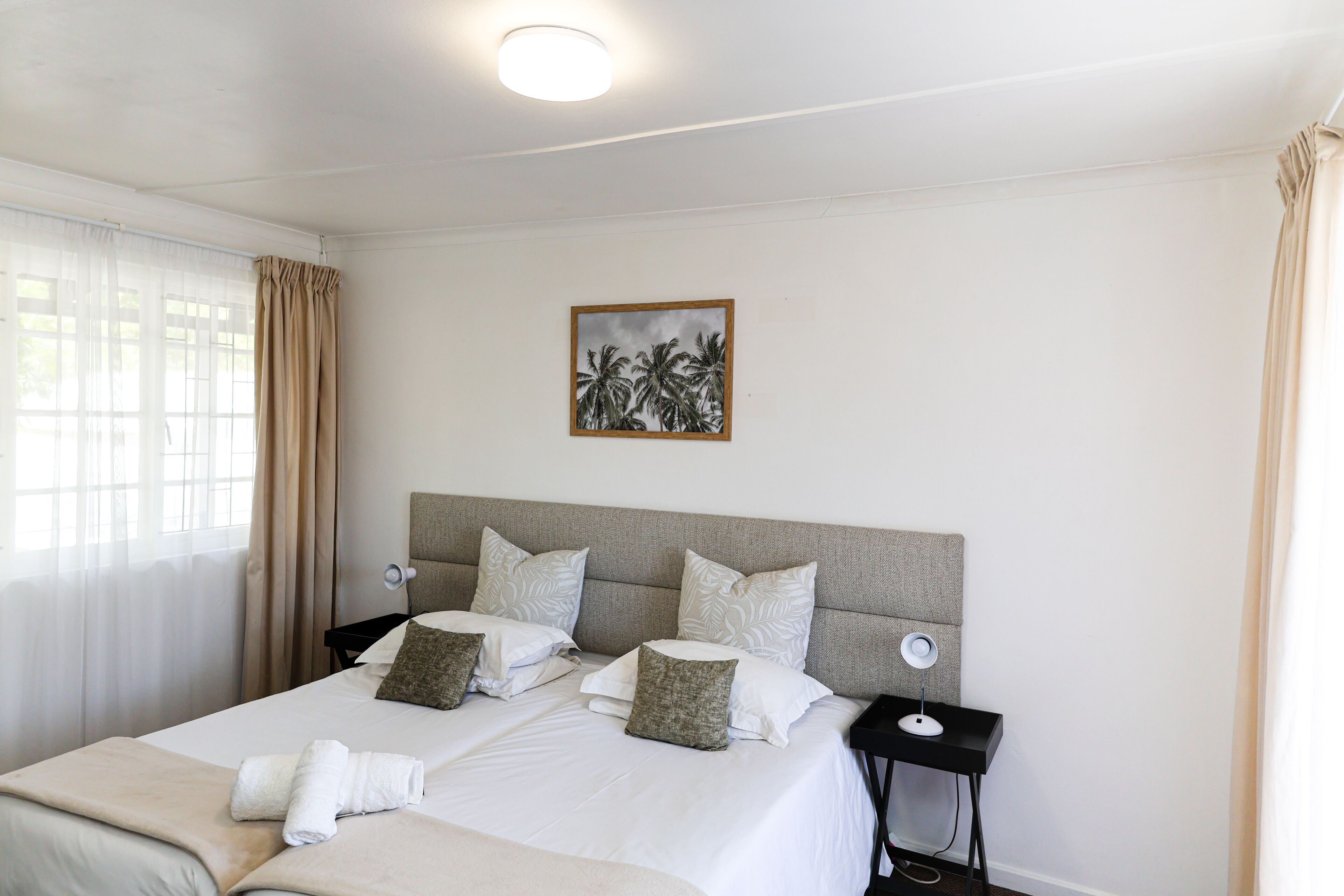 A neatly made bed with white linens and two pillows, a bedside table with a lamp, and a framed picture of palm trees on the wall.