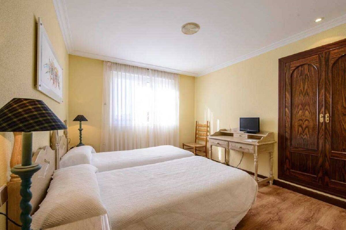 A hotel room with two beds, a desk with a television, a wooden wardrobe, and a window with curtains.