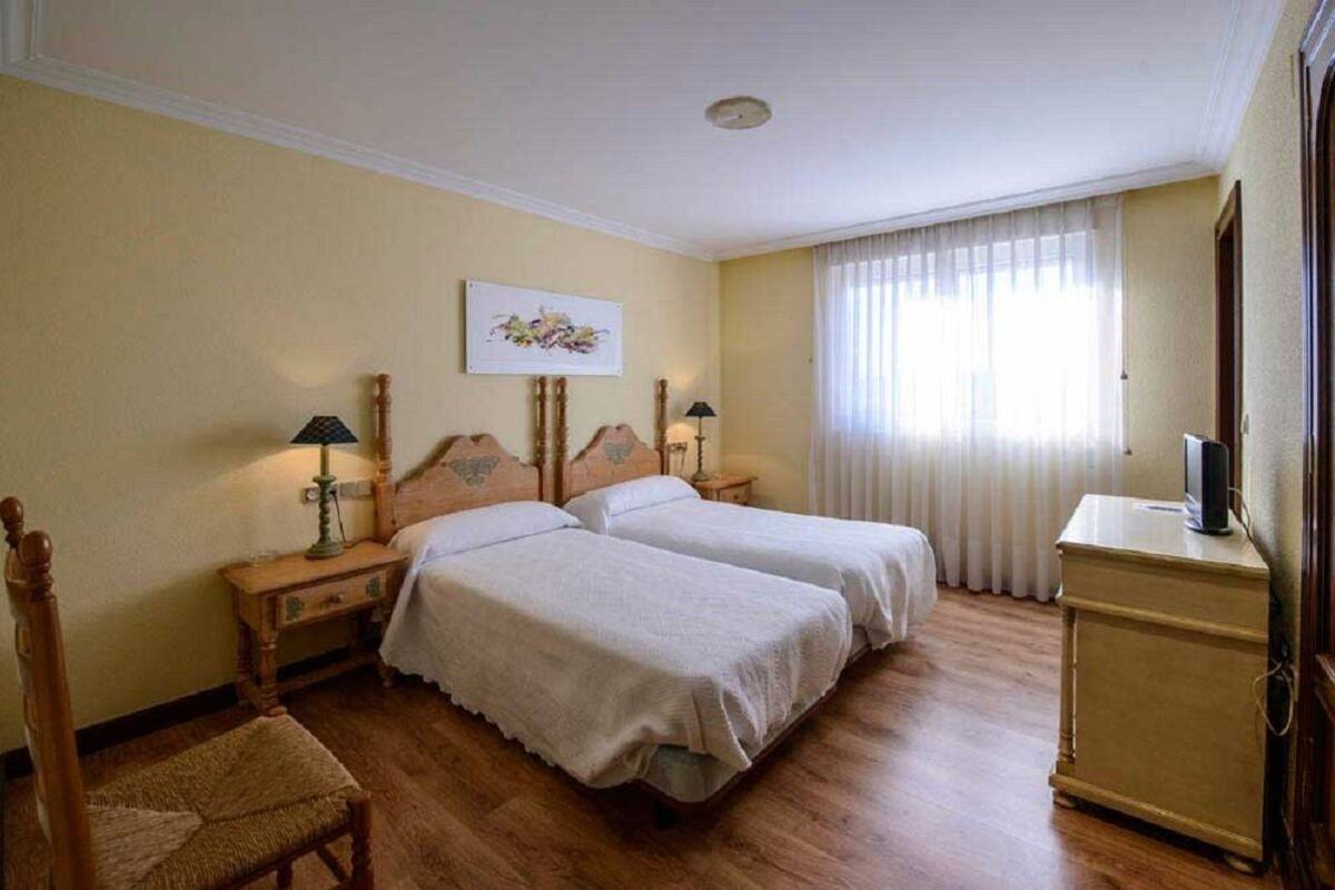 A hotel room with two single beds, wooden headboards, a small table with a lamp, a chair, a television on a cabinet, and a window with sheer curtains.
