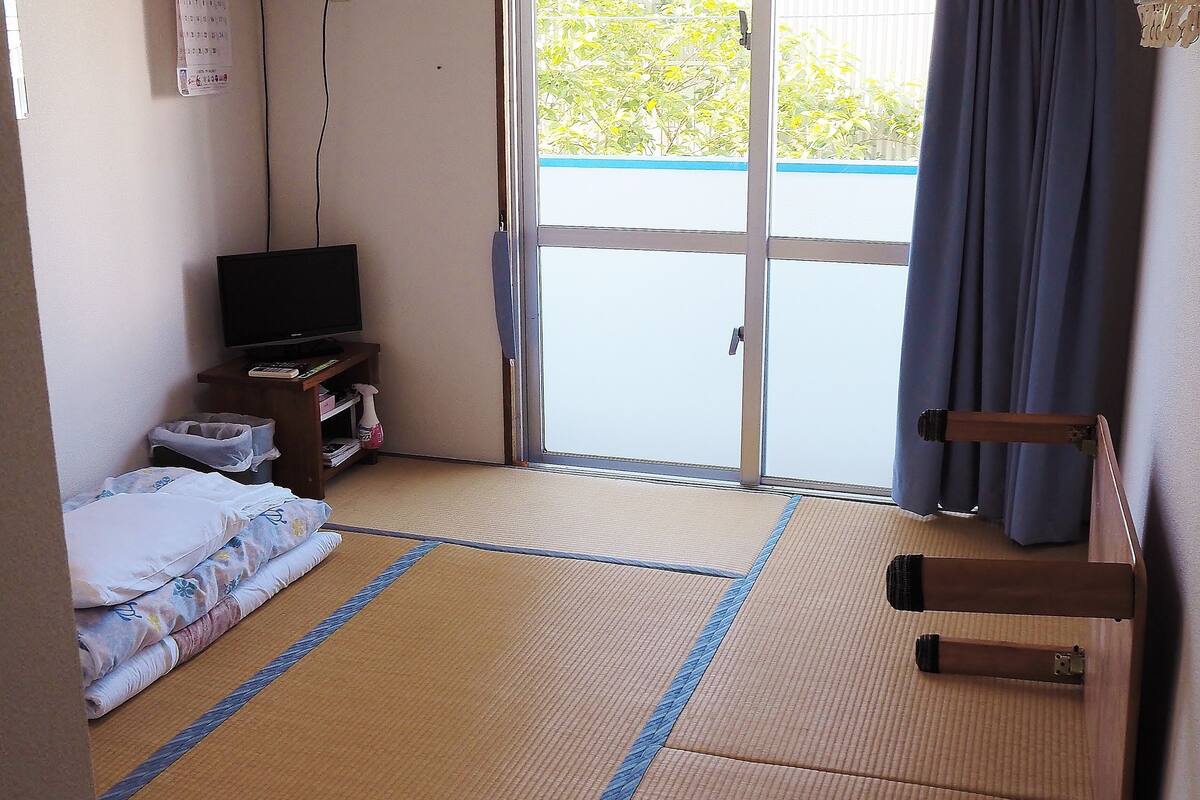 Une pièce avec un sol recouvert de tatamis, un téléviseur posé sur un meuble bas, un futon servant de lit, une porte-fenêtre coulissante donnant sur un espace extérieur et un ventilateur de plafond.