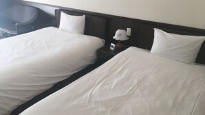 Dos camas individuales con sábanas blancas, un cabecero oscuro, una mesita de noche con lámpara y una silla.