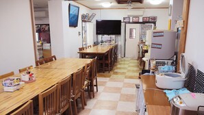 Un comedor con una mesa de madera larga y sillas, un televisor montado en la pared, y un refrigerador con varios artículos encima.