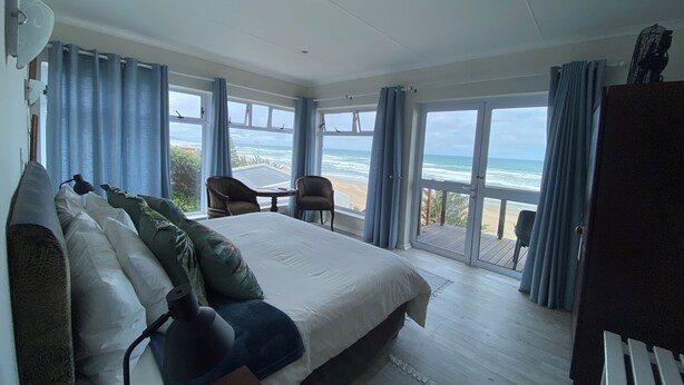 A bedroom with a large bed, two chairs, a small table, and a view of the ocean through large windows. - Sea Paradise (Wilderness)